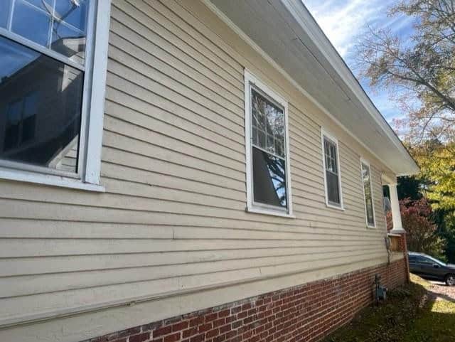 Side view of a cream-colored house with weathered siding, three white-framed windows, and a partial red-brick foundation. sunlight is visible and a tree reflects in one window.