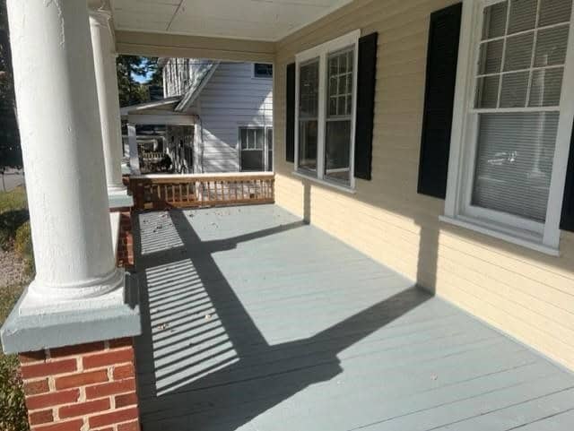 A sunlit front porch of a house with wooden flooring, white columns, and a wicker chair, casting sharp shadows across the surface. the house has grey siding and white-trimmed windows.