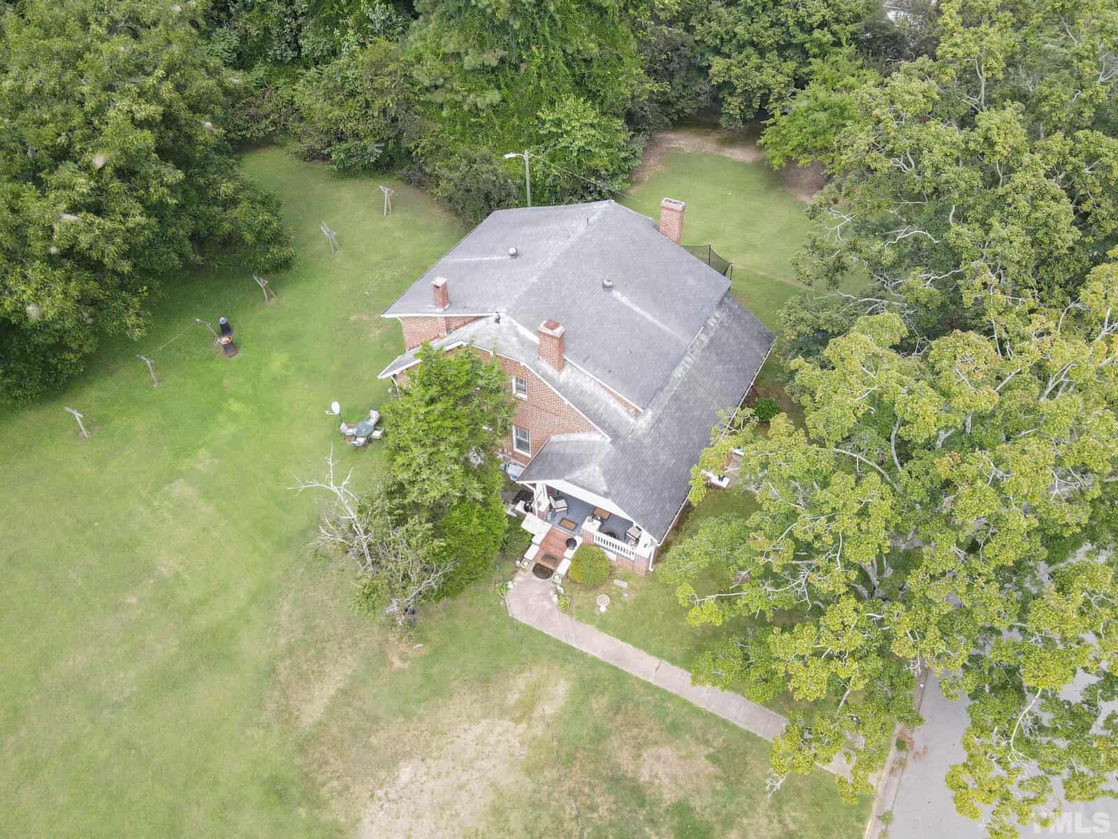 Aerial view of a two-story brick house with a shingle roof, surrounded by a green lawn with a few scattered trees.