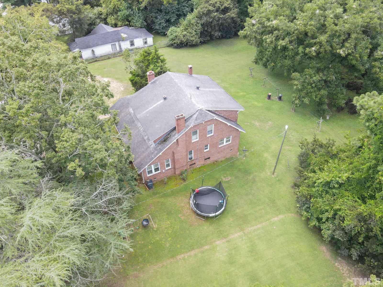 Aerial view of a red brick two-story house with a shingled roof, surrounded by a large green lawn with scattered trees, featuring an outdoor trampoline in the backyard.