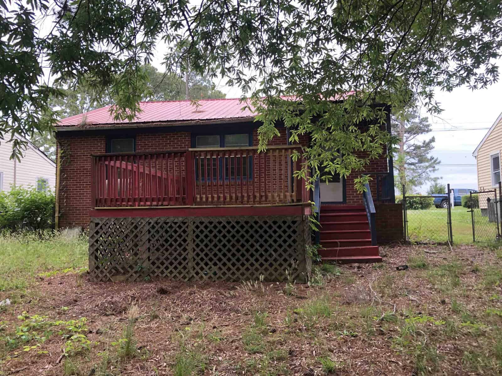 A small brick house with a red metal roof featuring a wooden front deck with lattice skirting and red steps, surrounded by a grassy area with trees.