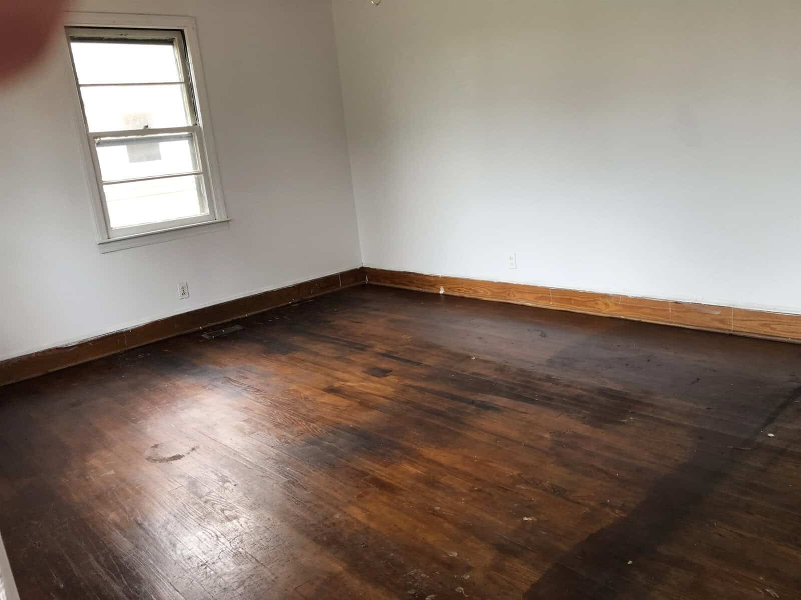 An empty room with bare hardwood floors and white walls, featuring a single window on the left wall.