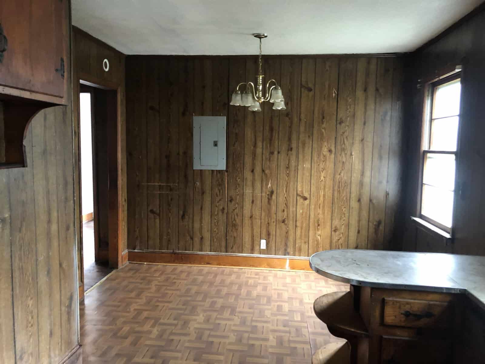 An empty room with wood paneled walls, a parquet floor, and a single light fixture hanging from the ceiling. there is a round table to the right and a window on the far wall providing natural light.