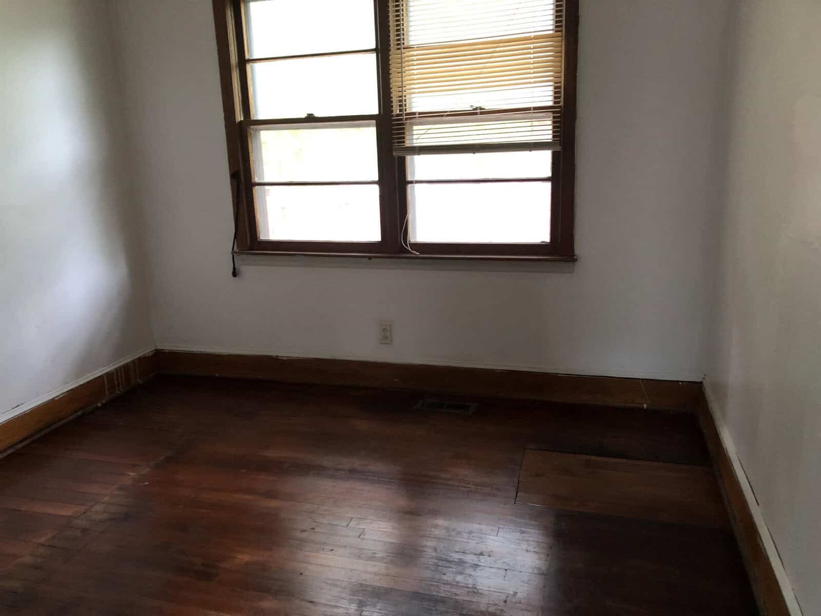 An empty room with hardwood floors, a double-hung window with a partially drawn blind, and white walls showing signs of wear.
