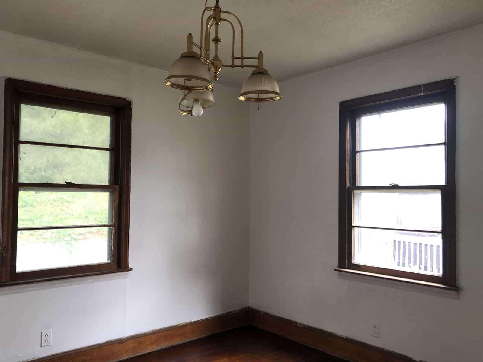 An empty room with white walls, two windows with a view of greenery, a chandelier, and hardwood flooring.