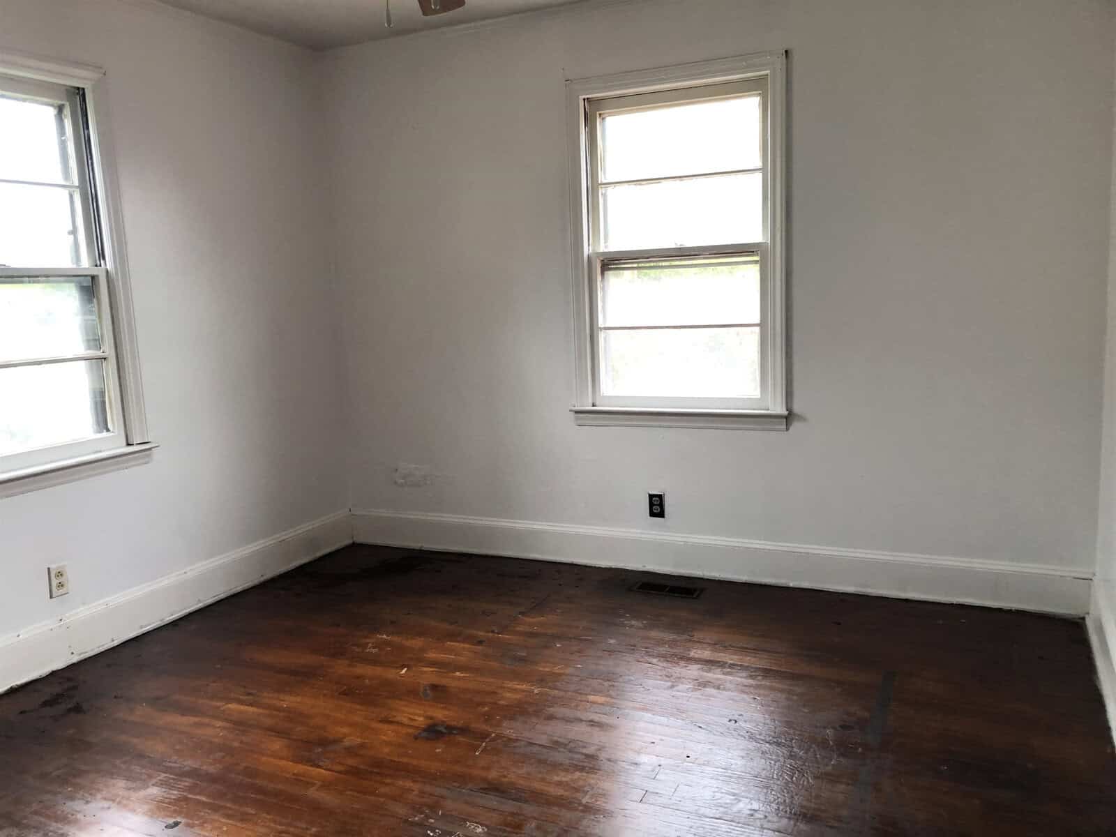 An empty room with white walls, two windows, and a hardwood floor showing signs of wear.