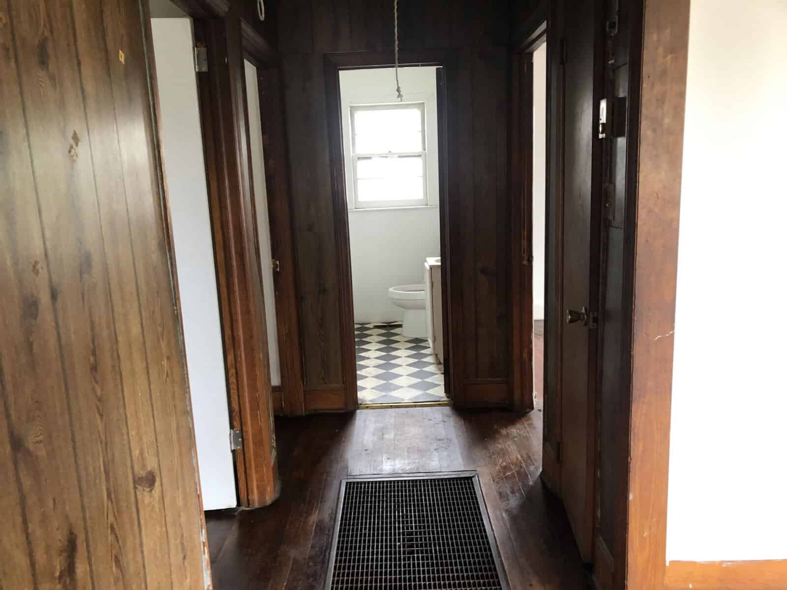 A narrow hallway with wooden paneling leading to a bathroom with a checkered floor, the bathroom door is open and the toilet is visible.
