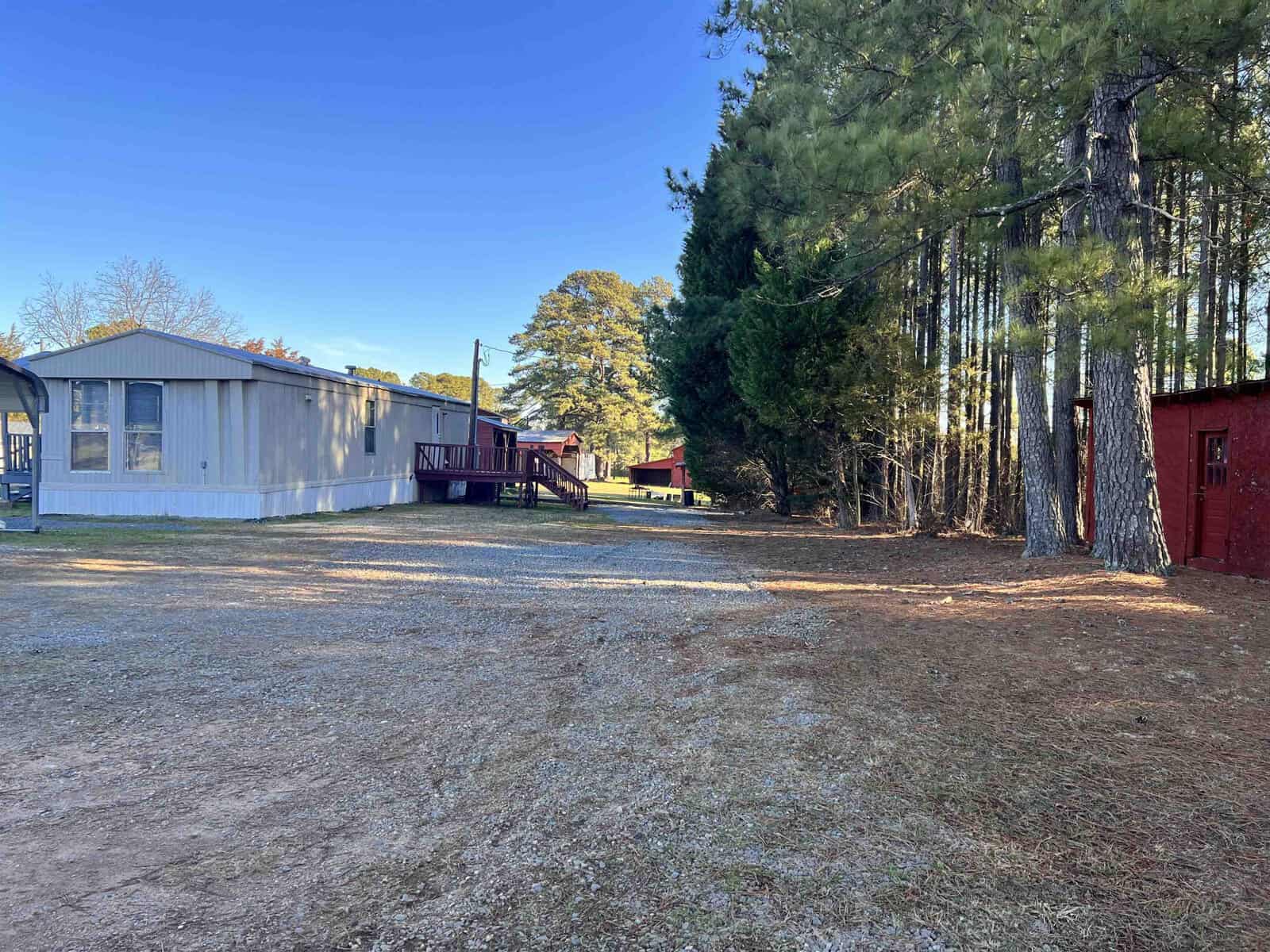 A mobile home with a small deck is situated on a plot of land with tall pine trees on the right. A gravel driveway leads to the home, and red buildings are visible in the background under a clear blue sky.