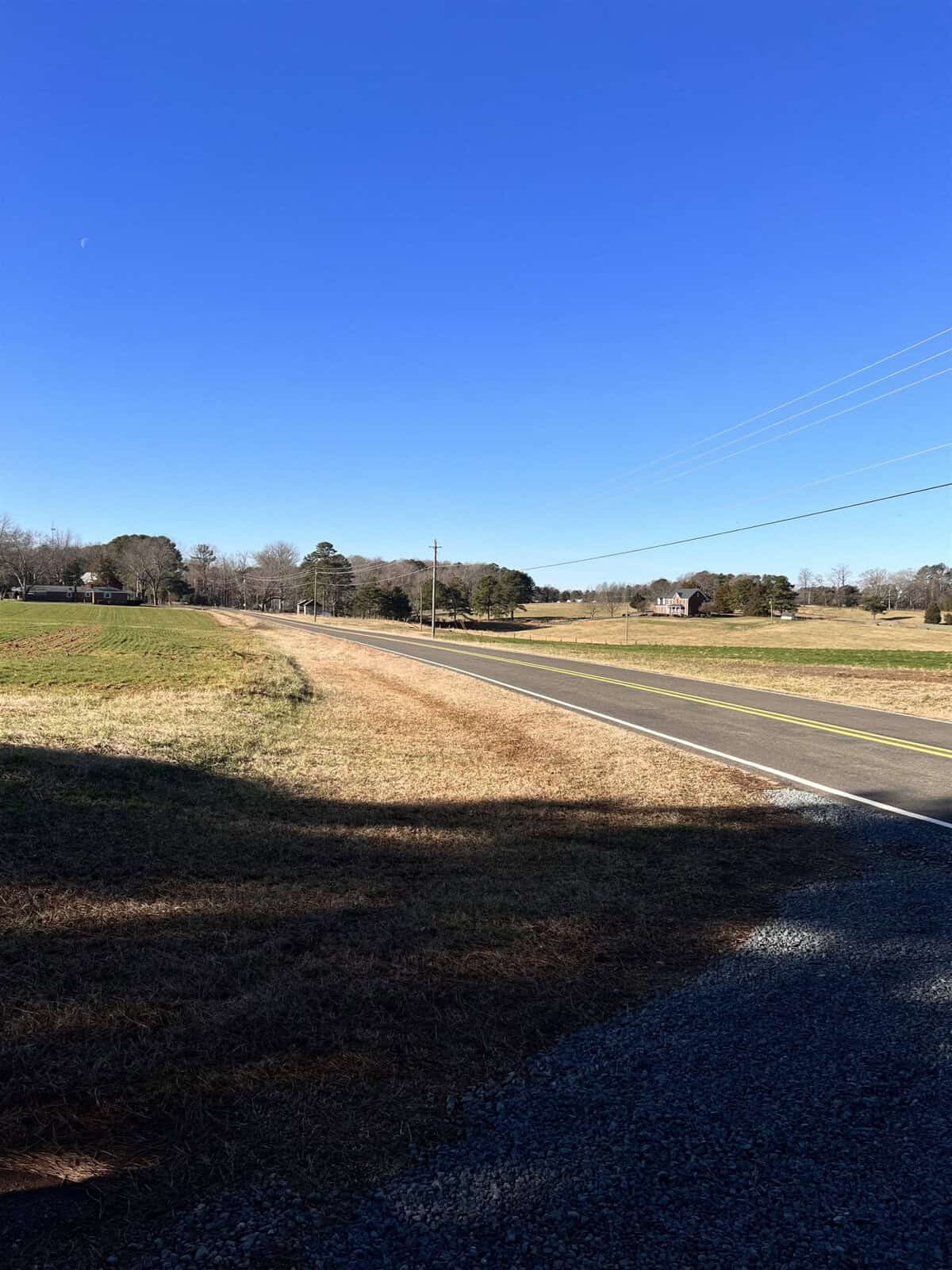 A rural landscape featuring a straight paved road running through grassy fields under a clear blue sky. Power lines follow the road, and a few trees and a distant house are visible in the background.