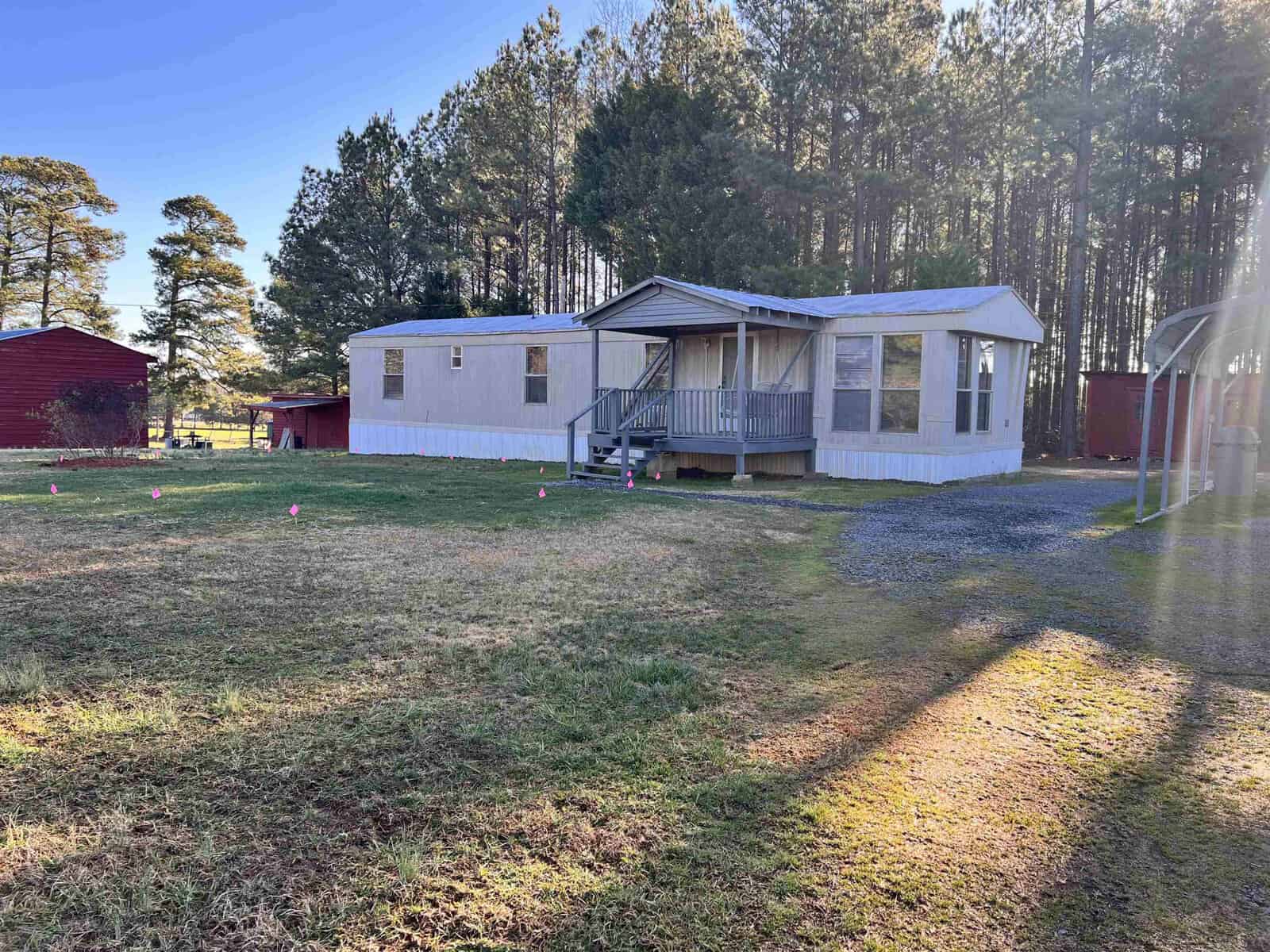 A mobile home with a wooden porch sits on a grassy lot. A red shed is visible on the left, and trees line the background. The gravel driveway is to the right, with sunlight casting long shadows on the grass.