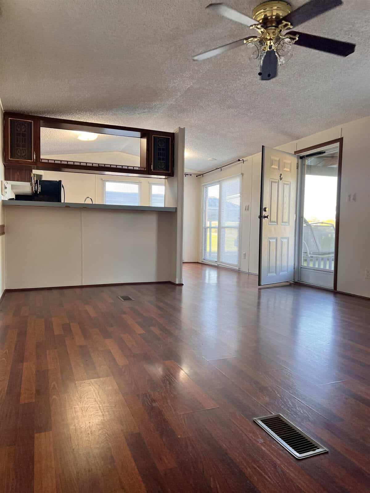 Spacious empty room with wooden floors, ceiling fan, and open kitchen area. Natural light streams through sliding glass doors, leading to a patio. The room features white walls and a decorative vent on the floor.