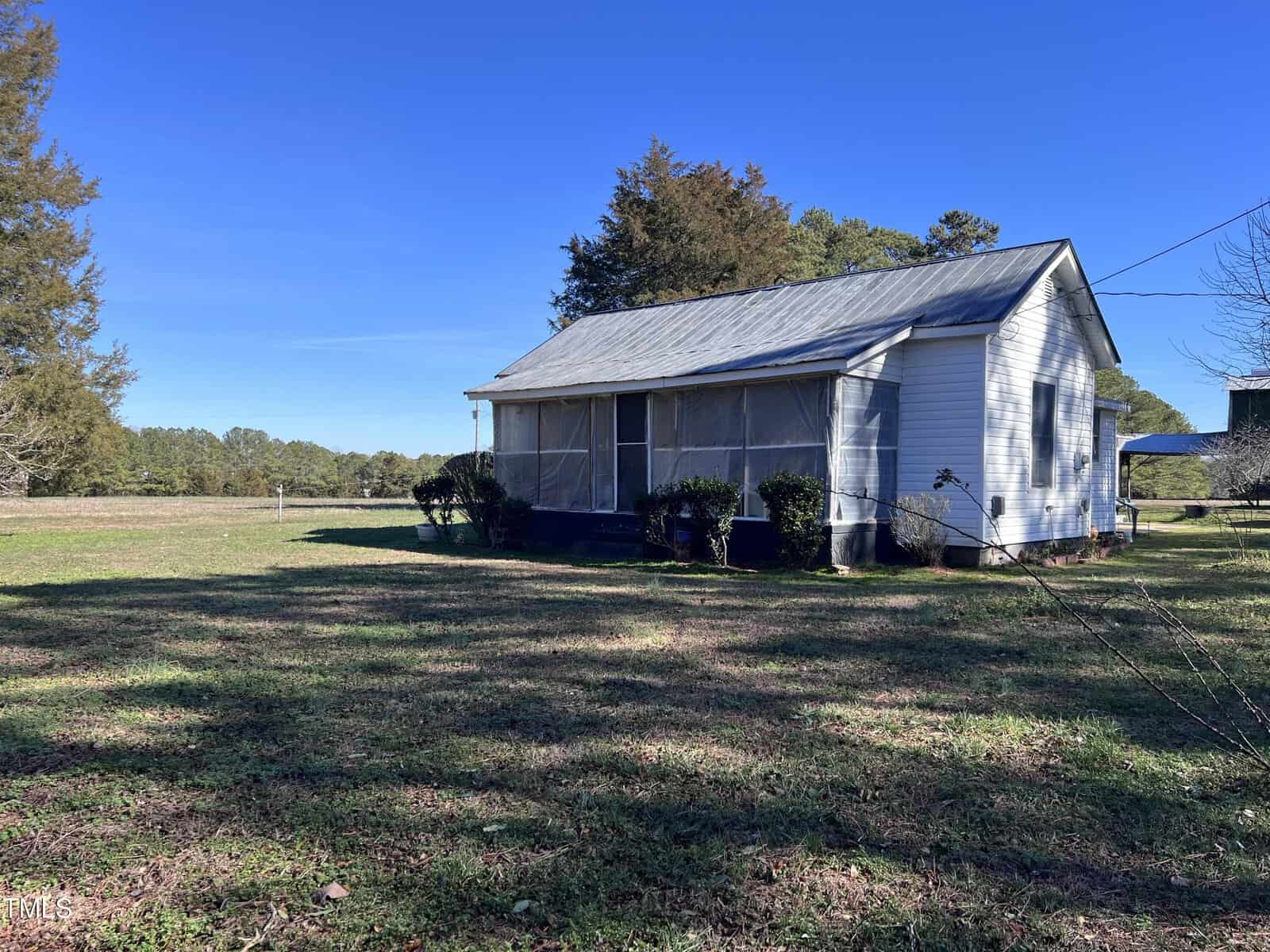 A small, white wooden house with a metal roof sits in a grassy field. The front porch is screened, and bushes line the front. Trees are in the background under a clear blue sky.