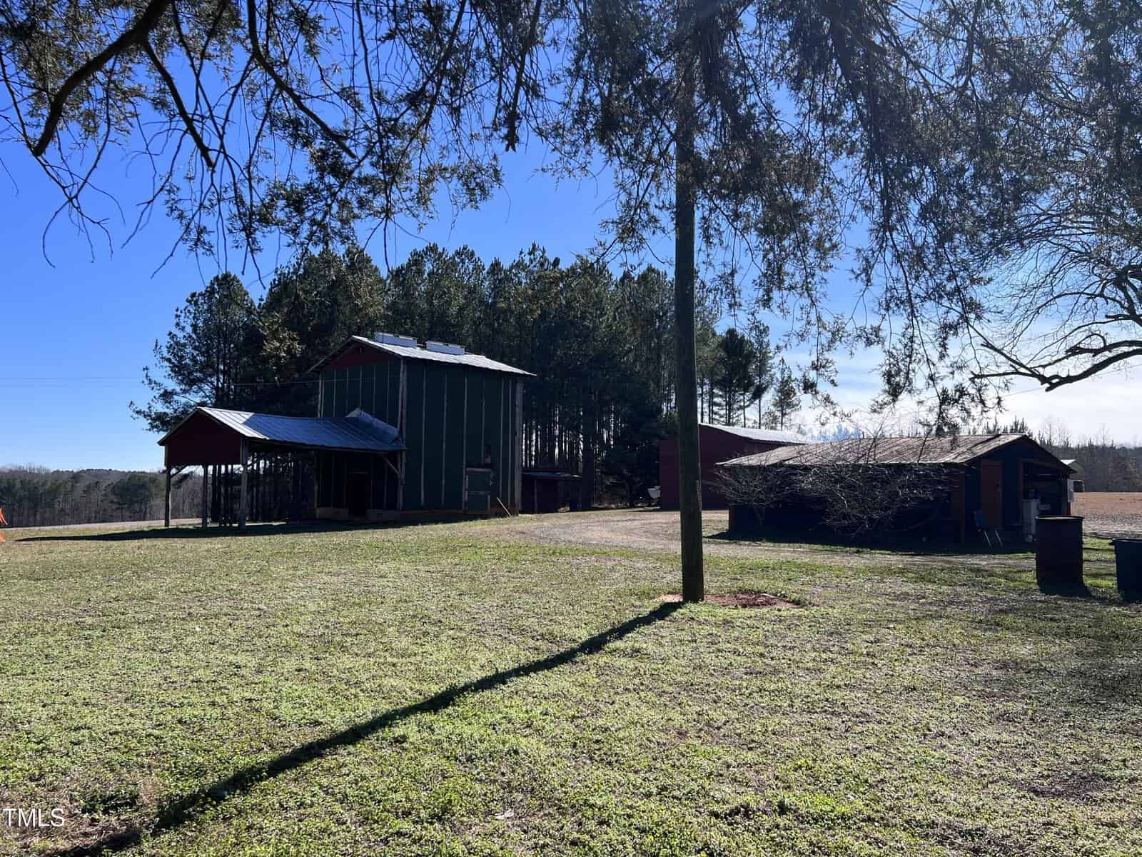 A rural scene with a green and red barn, surrounded by trees and an open field under a clear blue sky. Sunlight casts shadows on the ground and the area is calm and serene.