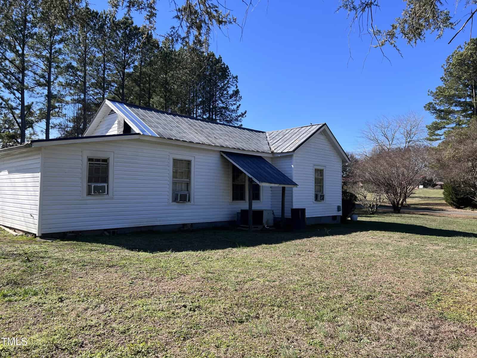 A small white house with a metal roof is surrounded by trees and a grassy yard. The house has several windows and an attached covered porch. The sky is clear and blue, and it's a sunny day.