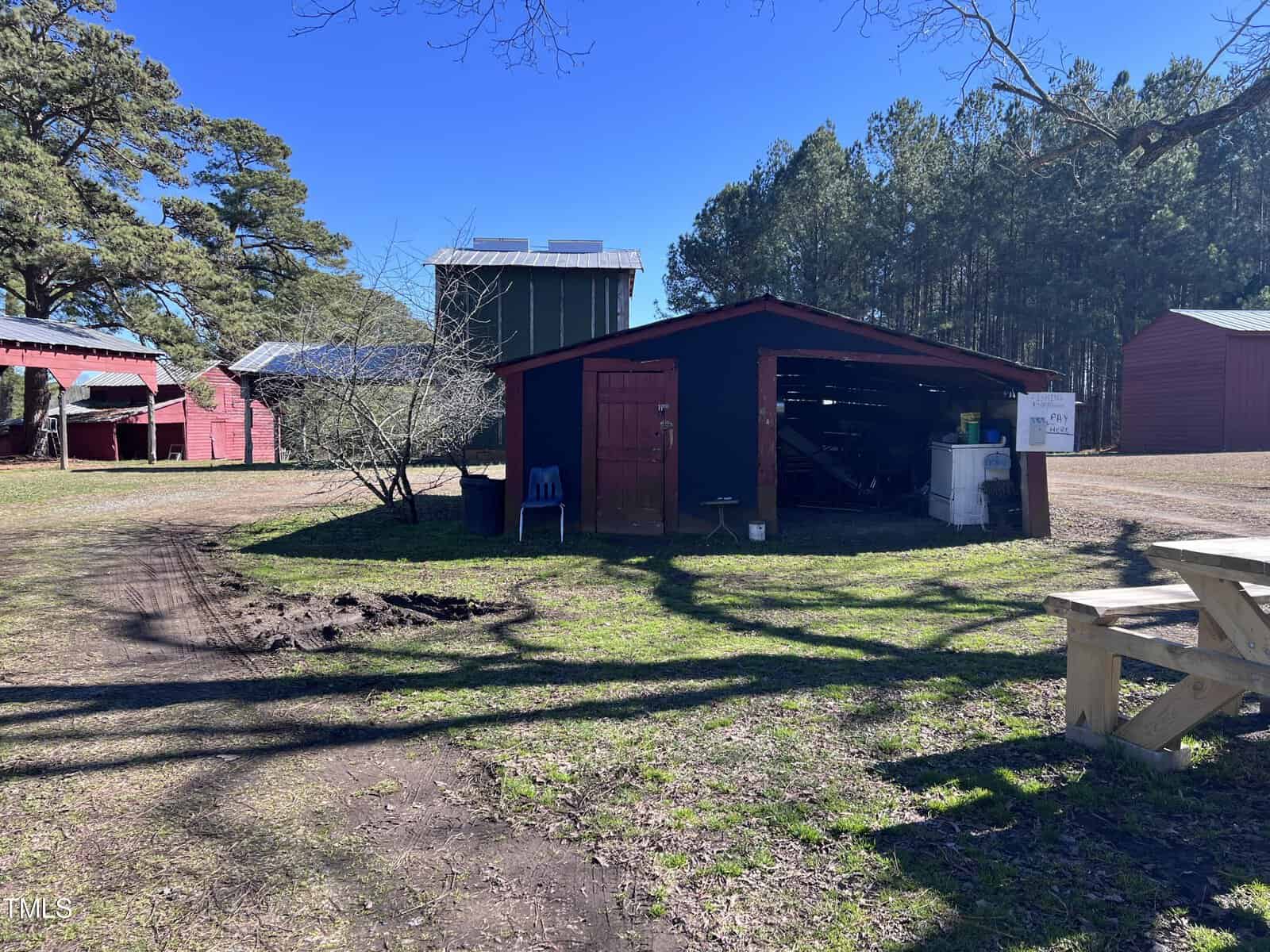 A rustic shed with an open door stands on a grassy patch, surrounded by tall trees. Nearby, a picnic table sits in the shade. A blue chair is visible beside the shed, and farm buildings are seen in the background under a clear blue sky.