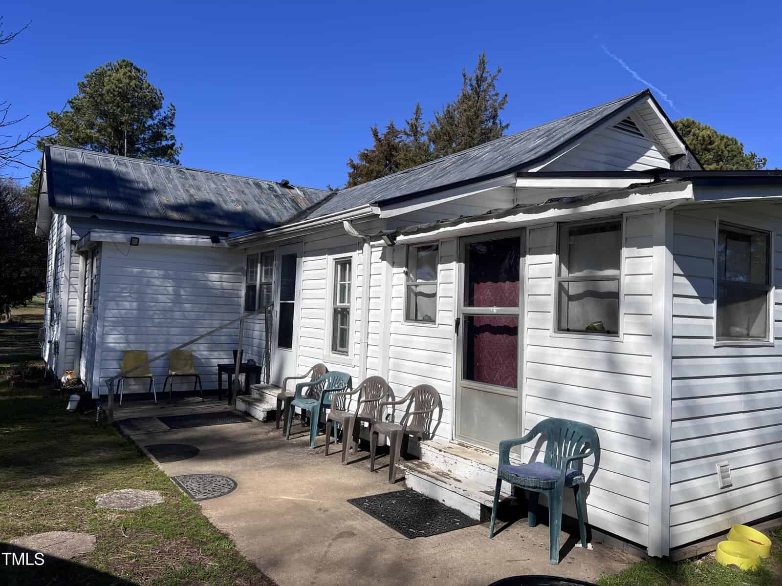 A small white house with a metal roof is shown. The porch is lined with several plastic chairs in various colors. There's a grassy yard and trees in the background under a clear blue sky.