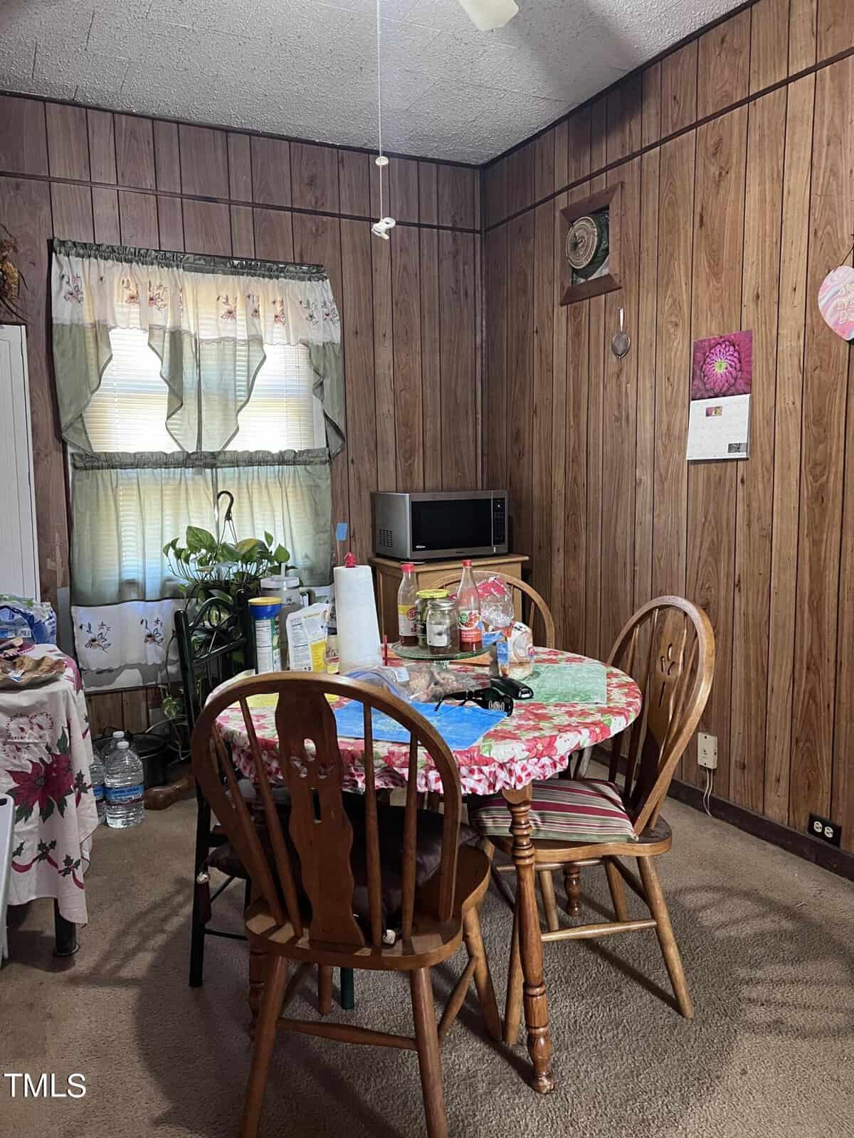 A small dining area with a round table covered by a floral tablecloth and four wooden chairs. The room has wood-paneled walls, a ceiling fan, a microwave on a shelf, and a window with floral curtains. Various items are on the table and nearby surfaces.