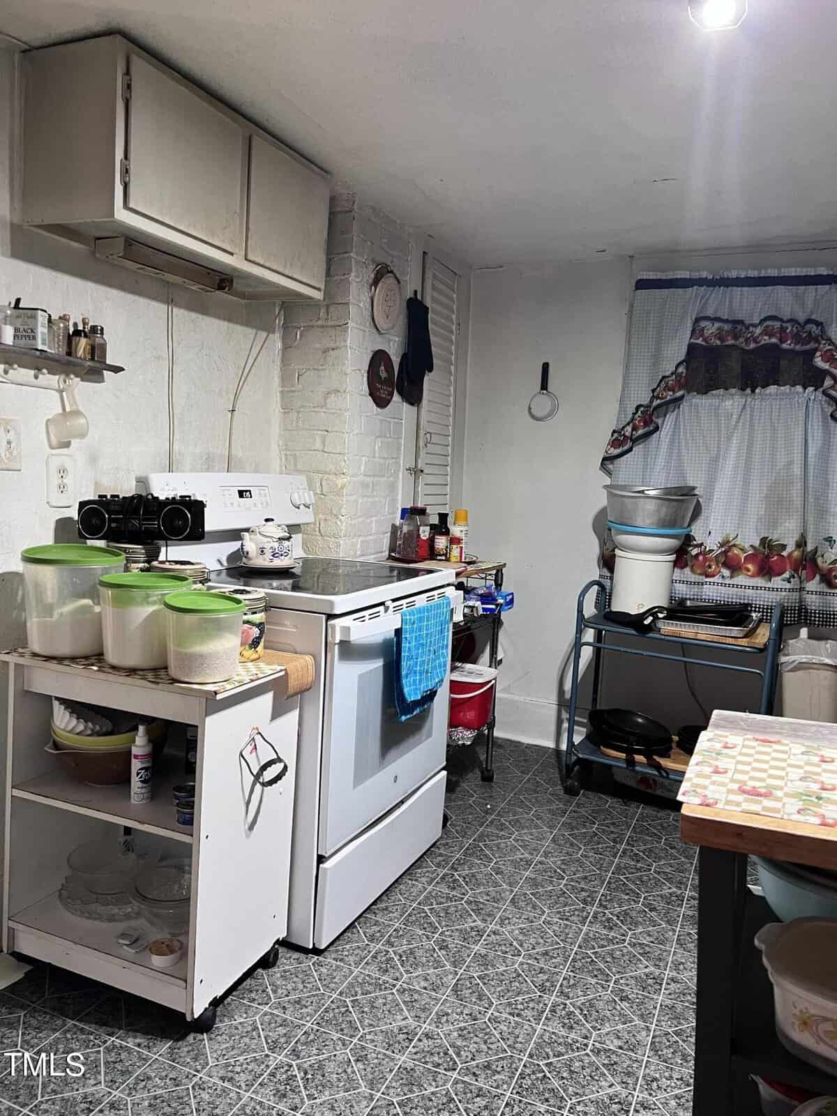 A small kitchen with a white stove, pots on top, and shelves with canned goods. A table with colorful patterned cloth holds kitchen items. A window with a ruffled curtain is visible, and the floor has a gray geometric pattern.