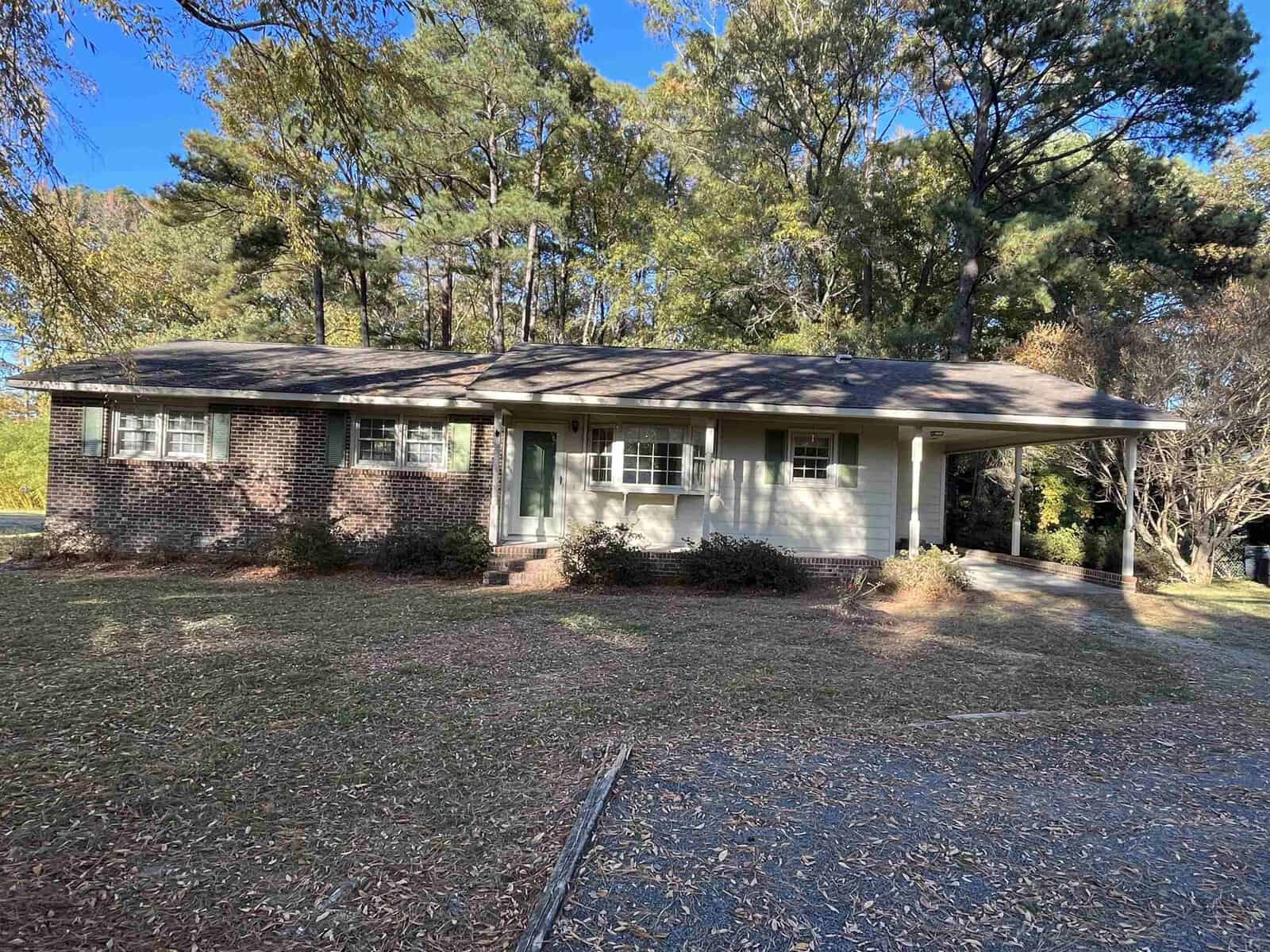 Nestled at 608 Lake Road in Creedmoor, this charming single-story brick house features a quaint front porch and attached carport. It's surrounded by tall trees and a lush grassy yard beneath a clear blue sky, offering a serene retreat.