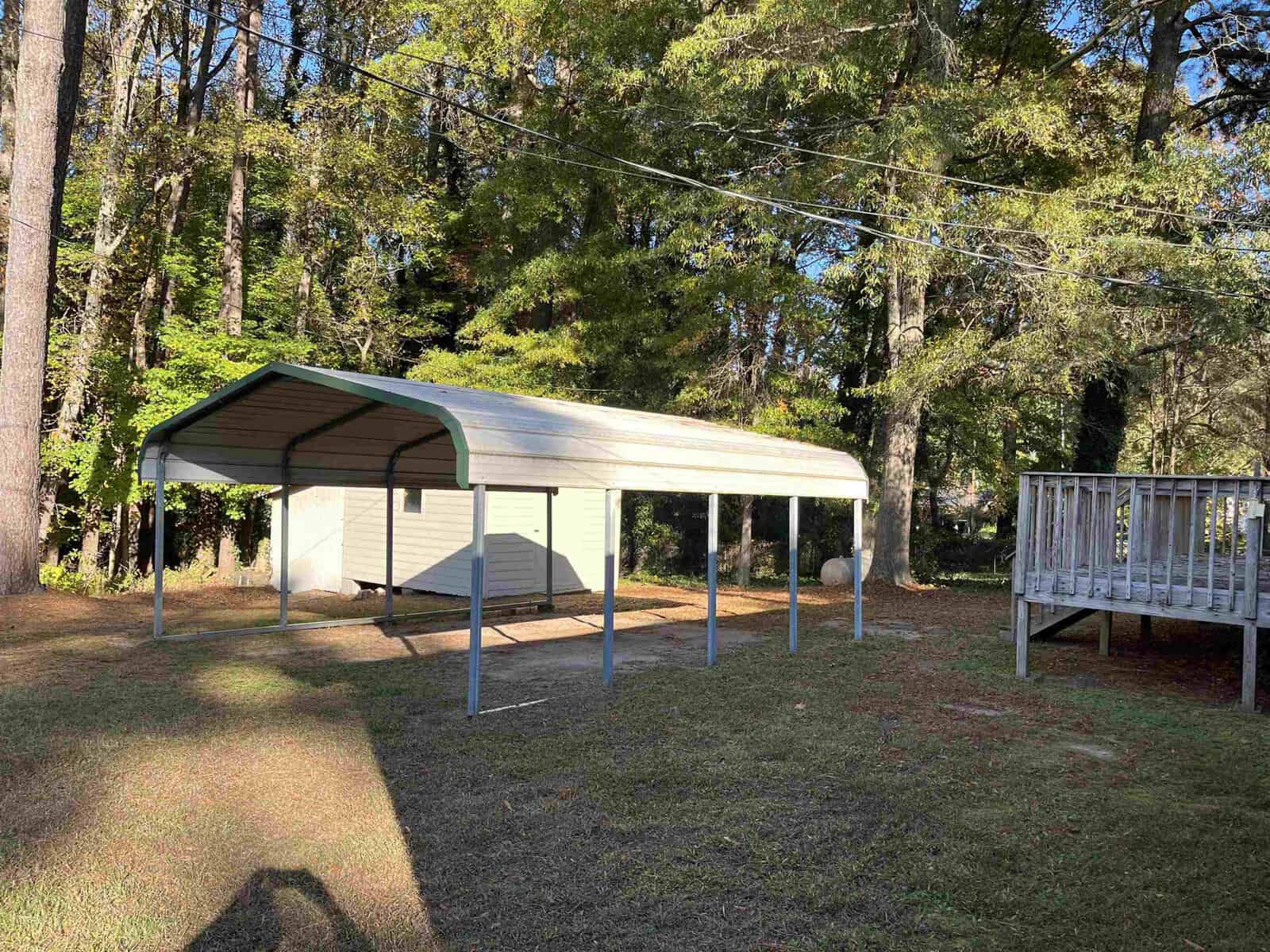 A metal carport stands on a grassy area next to a wooden deck. Tall trees surround the area, casting shade, and the sun is shining, creating dappled light on the grass.