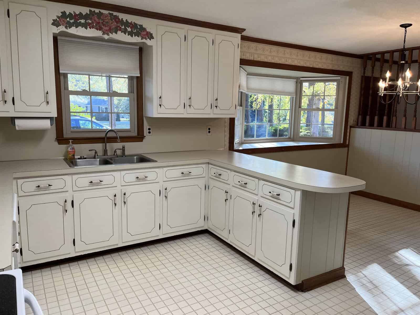 A vintage kitchen features white cabinetry, a large corner countertop, and a double sink. A window above the sink provides natural light, and a chandelier hangs in the adjoining dining area with a bay window and wooden accents.