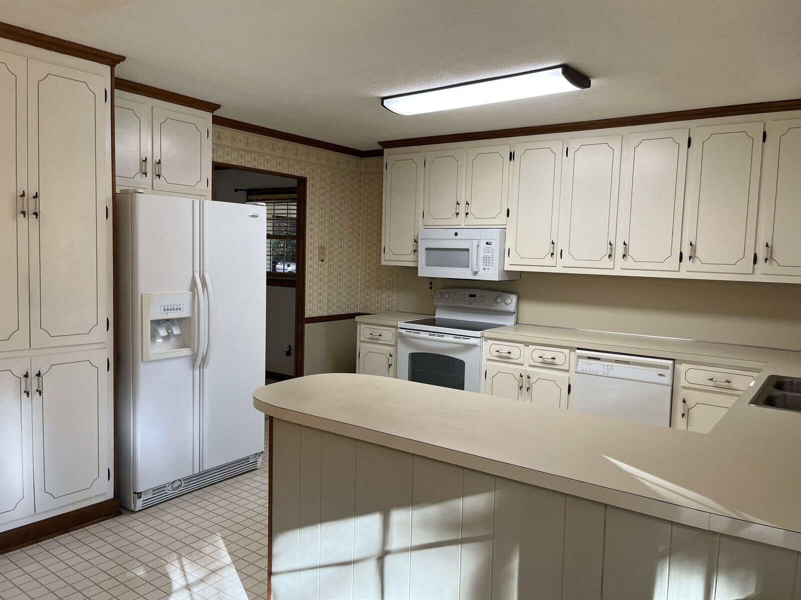 A kitchen with white cabinets, a white refrigerator, a white oven, a microwave, and a dishwasher. A curved countertop is in the foreground, and a fluorescent light fixture is on the ceiling. The floor has square tiles.