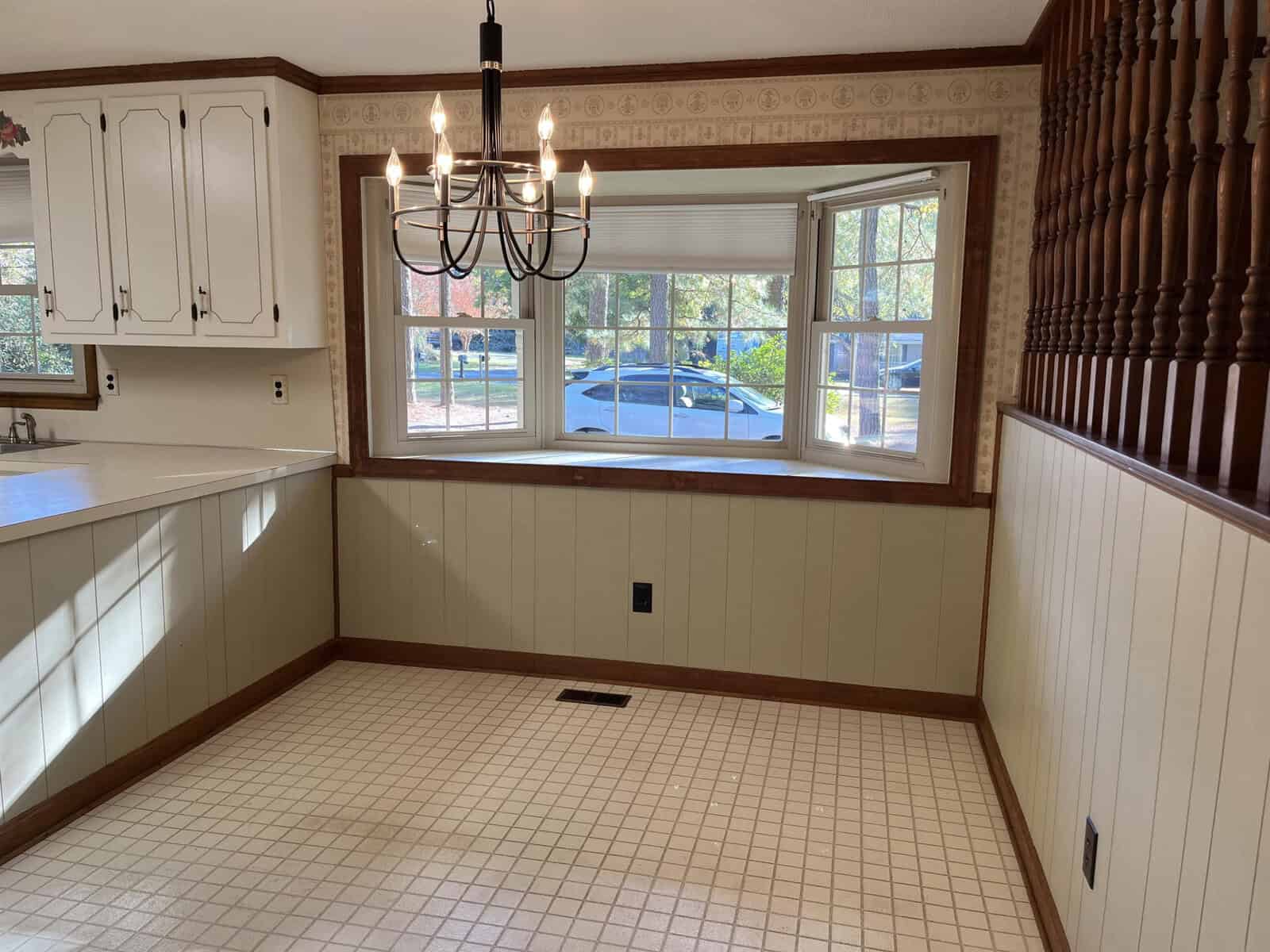 A bright dining area with white paneled walls and a window overlooking a parked car outside. A black chandelier hangs from the ceiling. The room has white cabinets and a light-colored tiled floor.