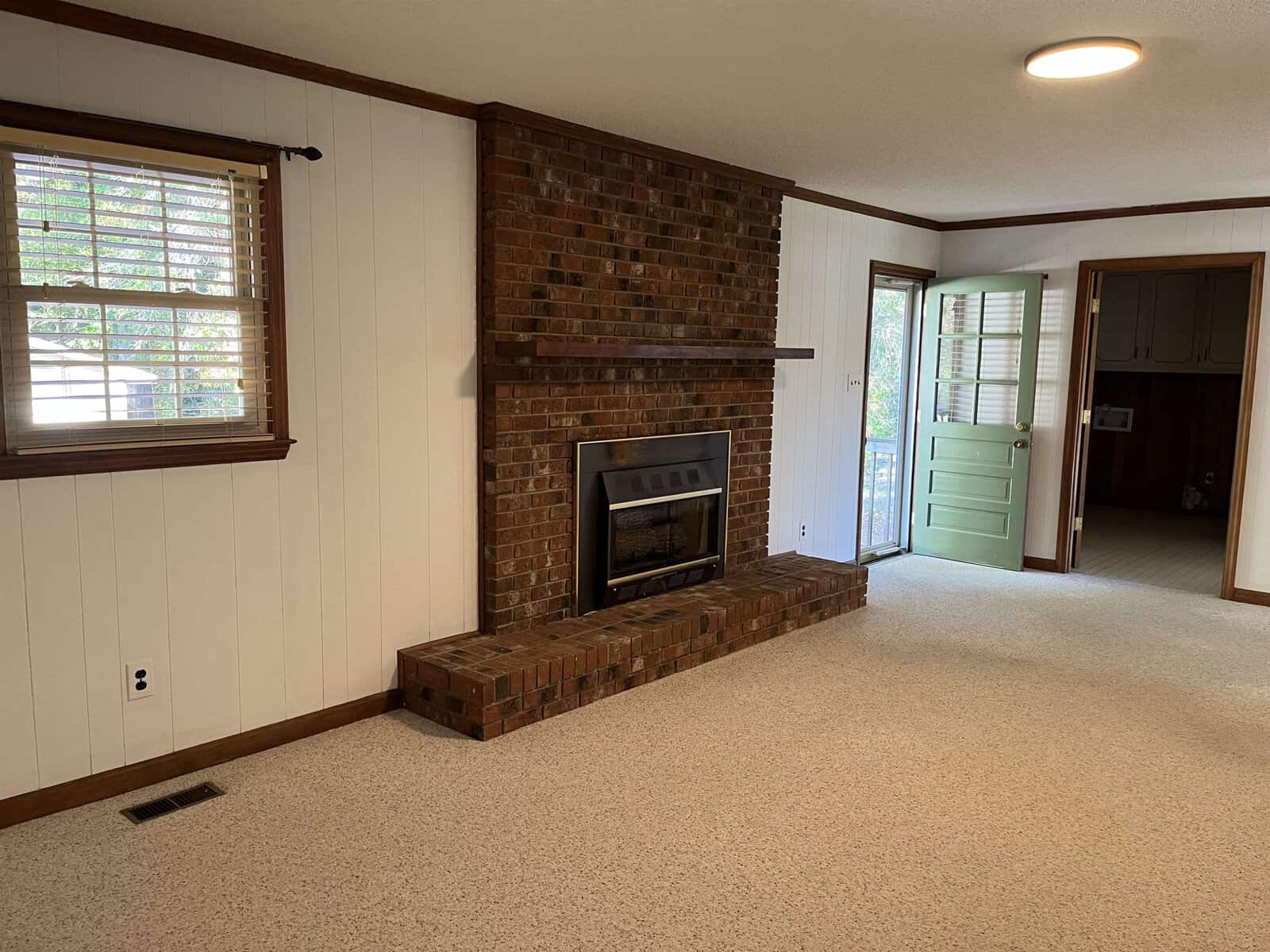 A cozy living room with a brick fireplace centered on a white paneled wall. The room has beige carpet, a window with wooden blinds, and a green door leading outside. There is an open doorway to another room.