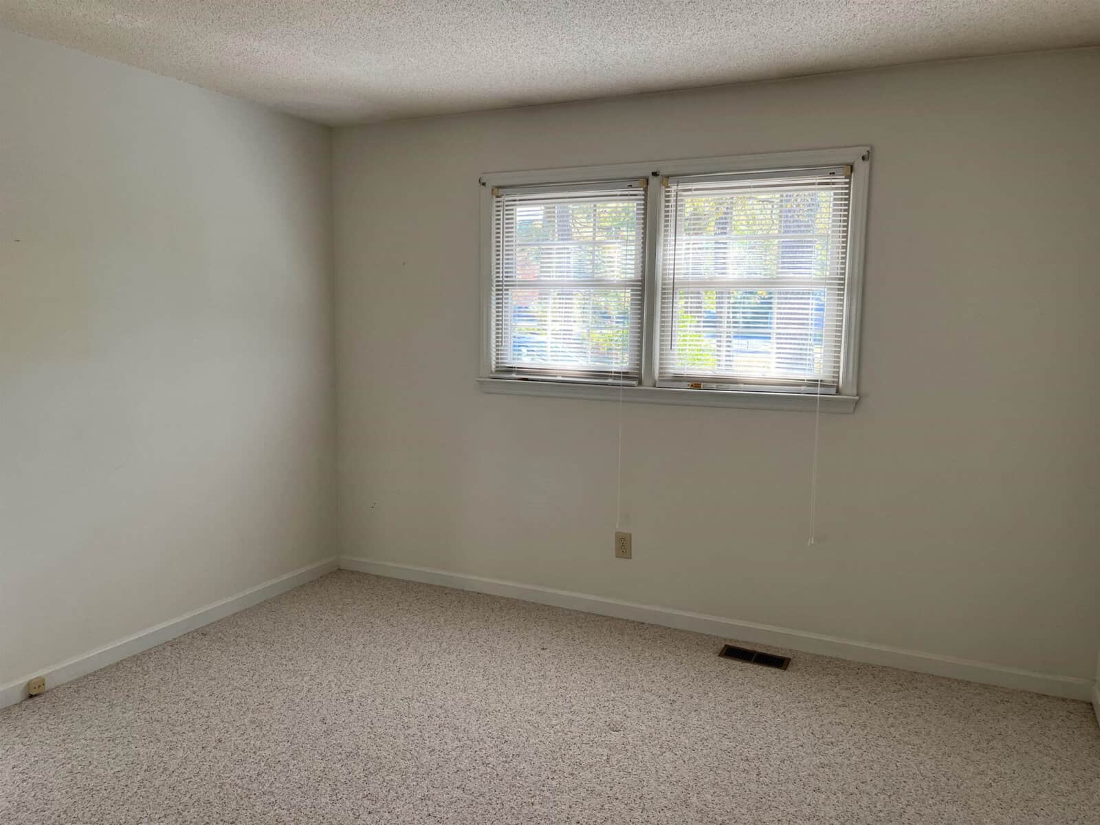 An empty room with white walls and beige carpet. Two windows with blinds let in natural light. A single electrical outlet is visible on the wall, and a floor vent is near the baseboard. The room has a simple and clean appearance.