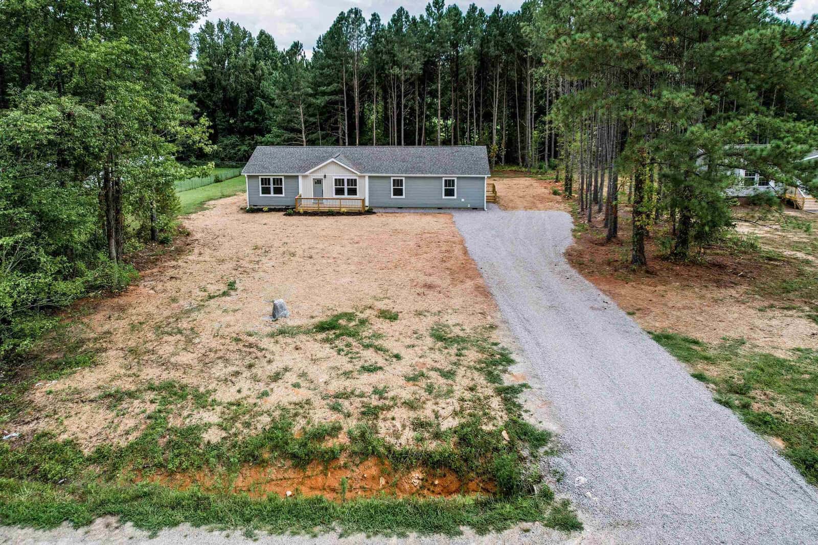 A single-story house with a gray roof and beige siding, featuring an extended driveway, is situated in a clearing surrounded by tall pine trees. the terrain appears dry with patches of grass, and a lone figure stands in the open area in front of the house.