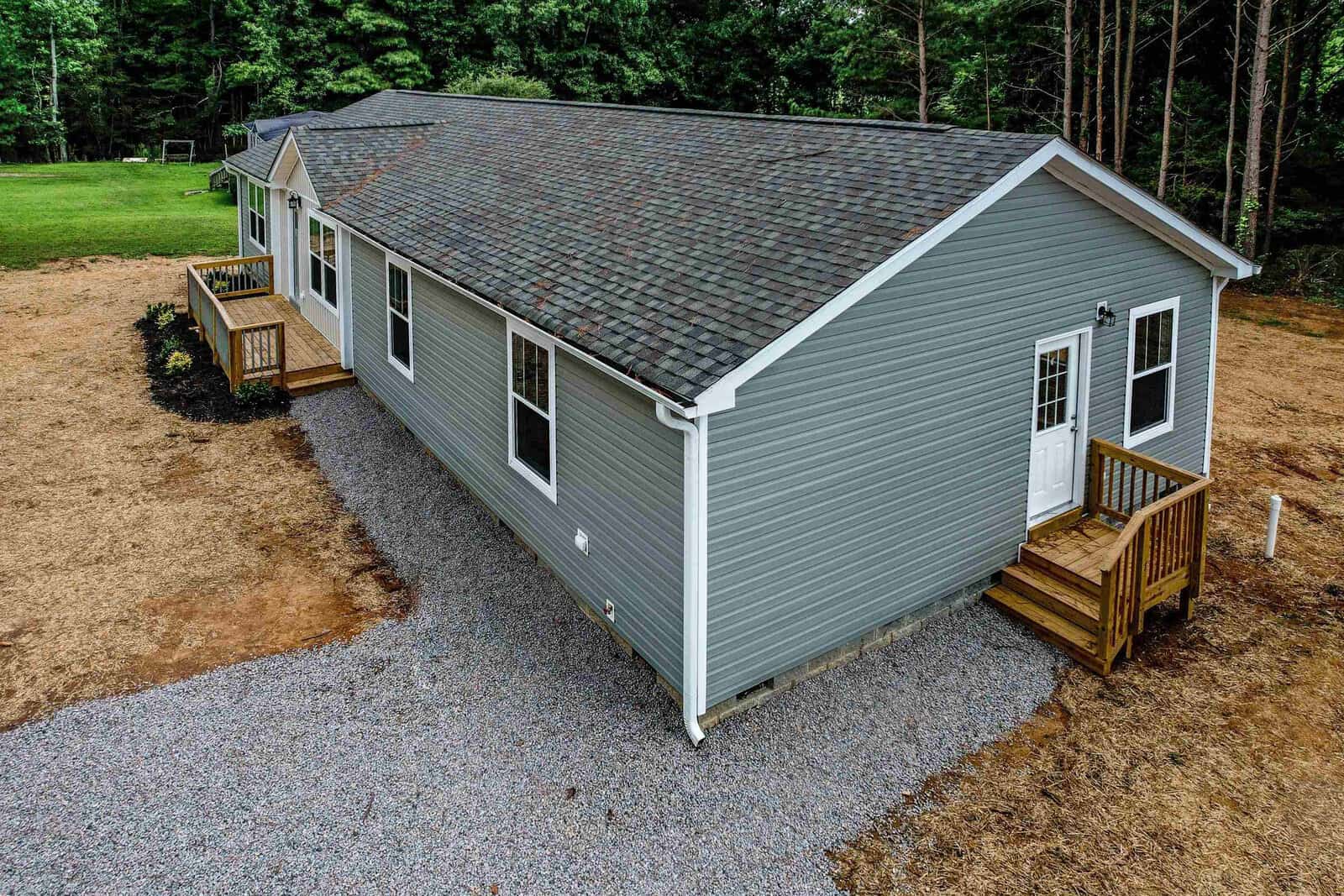 Aerial view of a single-story gray house with a pitched roof, featuring wooden front and side steps, situated on a landscaped lot with a gravel surround.