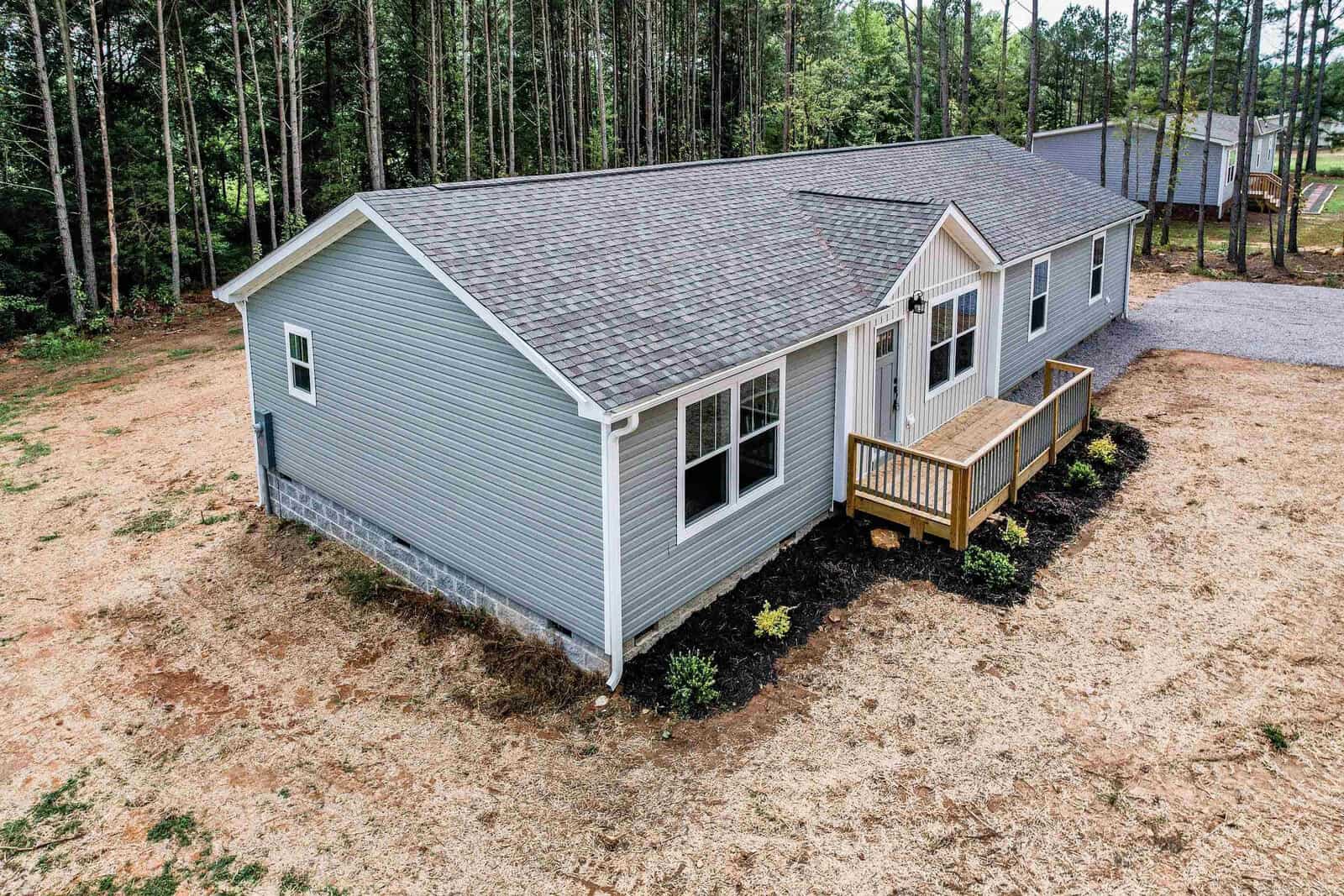 Aerial view of a single-story suburban house with gray siding and a shingle roof, featuring a small front porch and surrounded by a sparsely grassed yard with a backdrop of trees.
