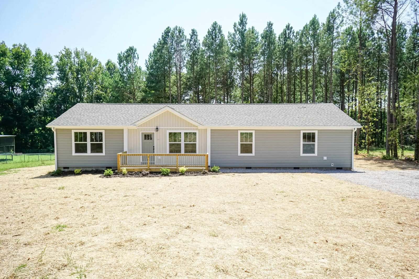 A single-story gray house with white trim and a small front porch, situated in a cleared area with sparse grass and surrounded by tall pine trees.