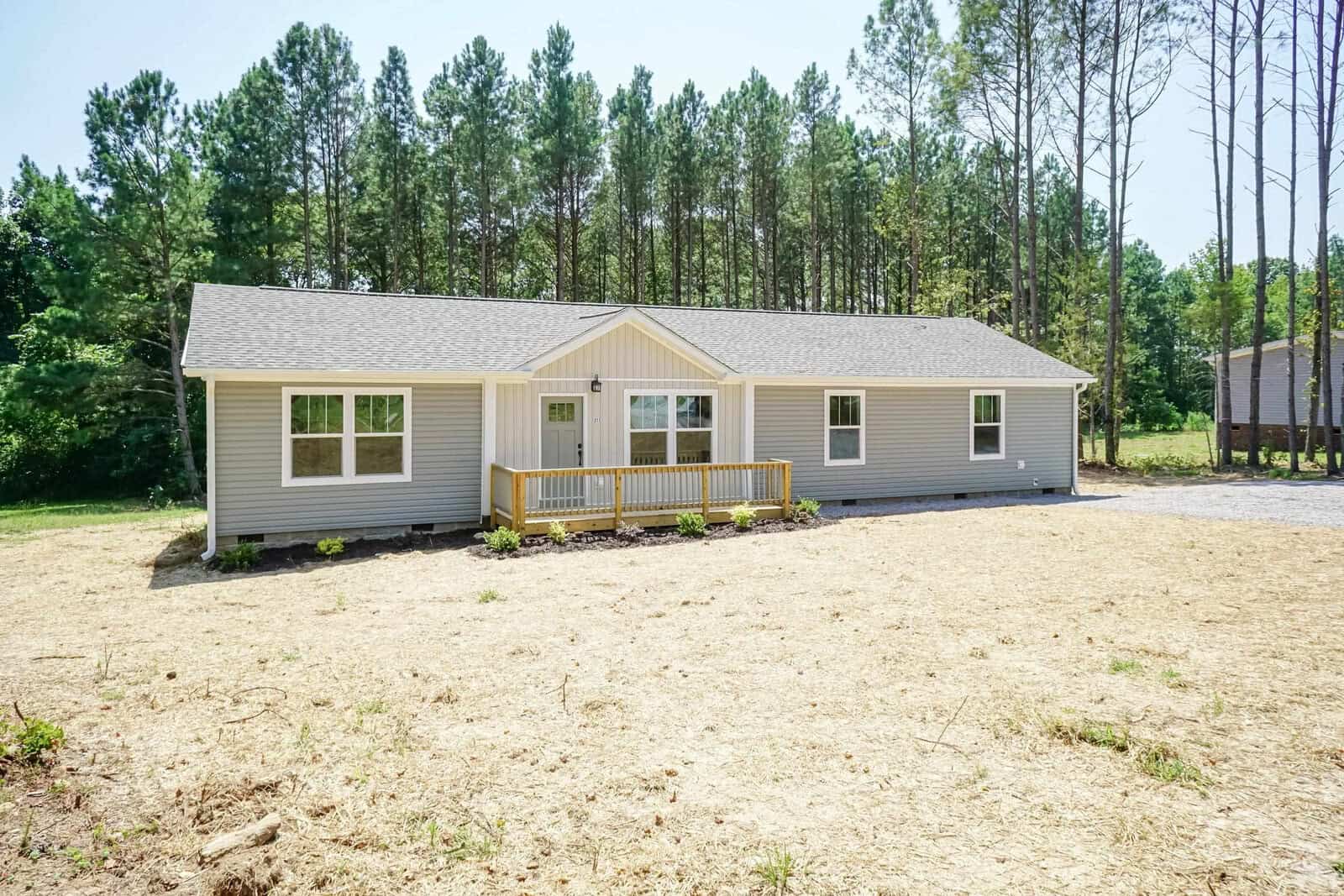 A single-story gray house with white trim and a small front porch, surrounded by a sparse lawn with a backdrop of tall pine trees.