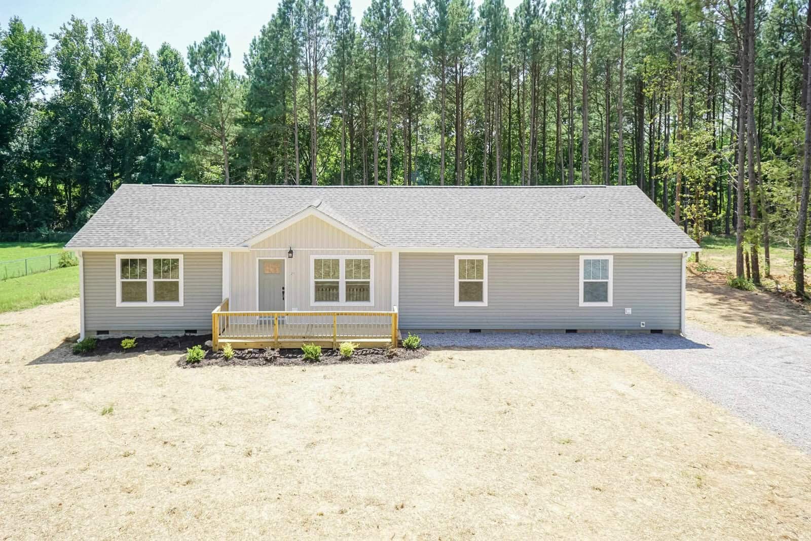 A single-story gray house with white trim and a yellow front door, featuring a gravel driveway and situated in a clearing surrounded by trees.