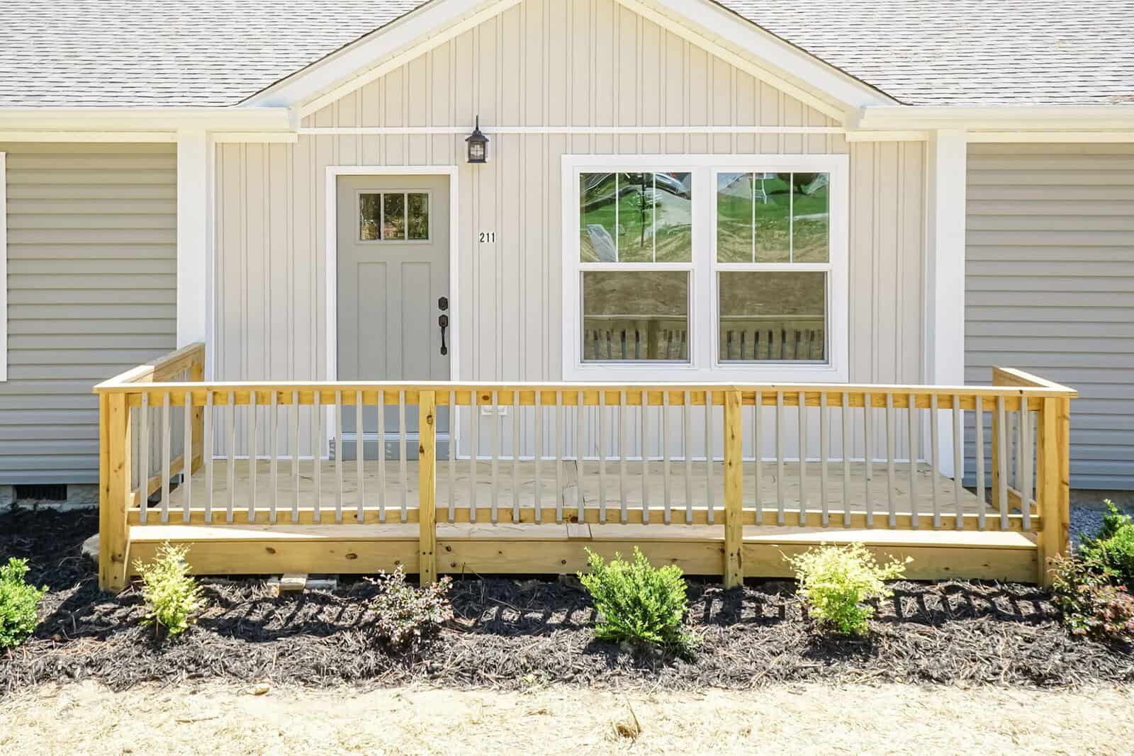 A new single-story house with beige vinyl siding, featuring a small wooden front porch with railings, a white front door, and a double window. there are young shrubs planted in the mulched bed in front of the porch.
