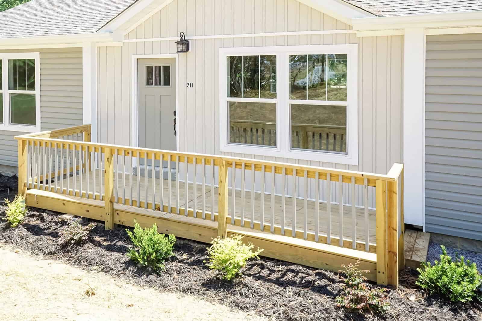 A newly constructed home with beige vinyl siding, featuring a small wooden front deck with railings, flanked by young shrubbery and covered with mulch.