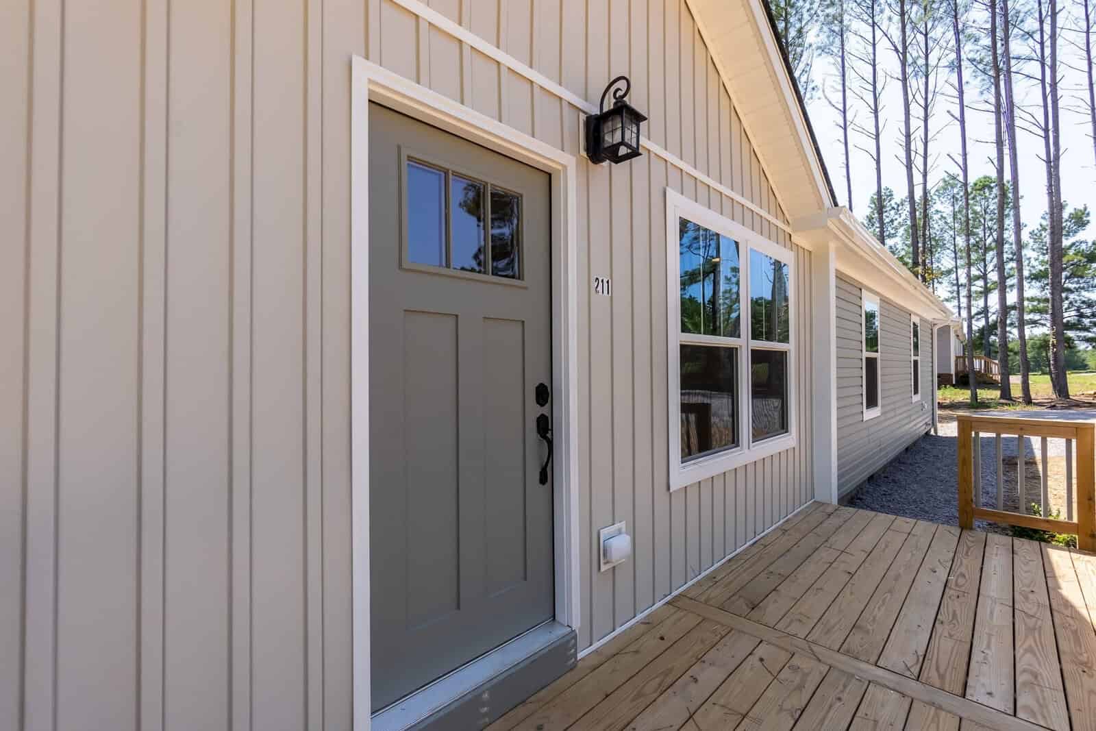 A new beige siding house with wooden porch, showing a gray front door with a window, next to a black outdoor light fixture, under clear skies.