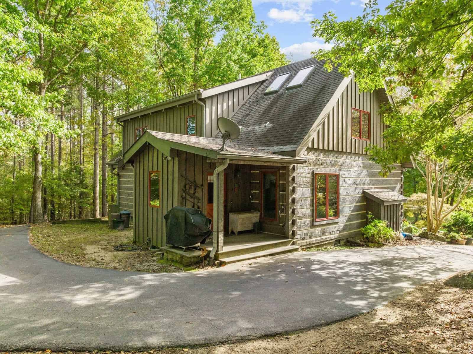 A rustic, two-story cabin made of wood planks with red-trimmed windows sits among dense, leafy trees. The cabin features a small, covered porch with a satellite dish on the roof and a paved driveway curving to its entrance. The setting is serene and green.