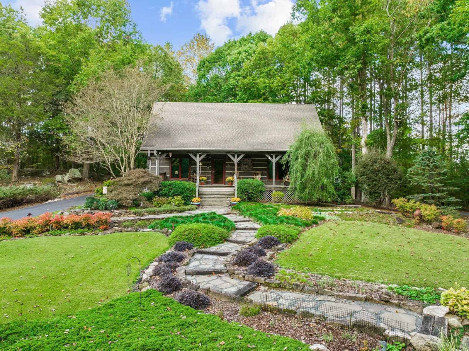 A rustic cabin sits surrounded by vibrant greenery and landscaped gardens. A stone pathway leads through manicured lawns, bordered by colorful flowers and shrubs, up to the cabin's front porch. Tall trees provide a serene, natural backdrop.