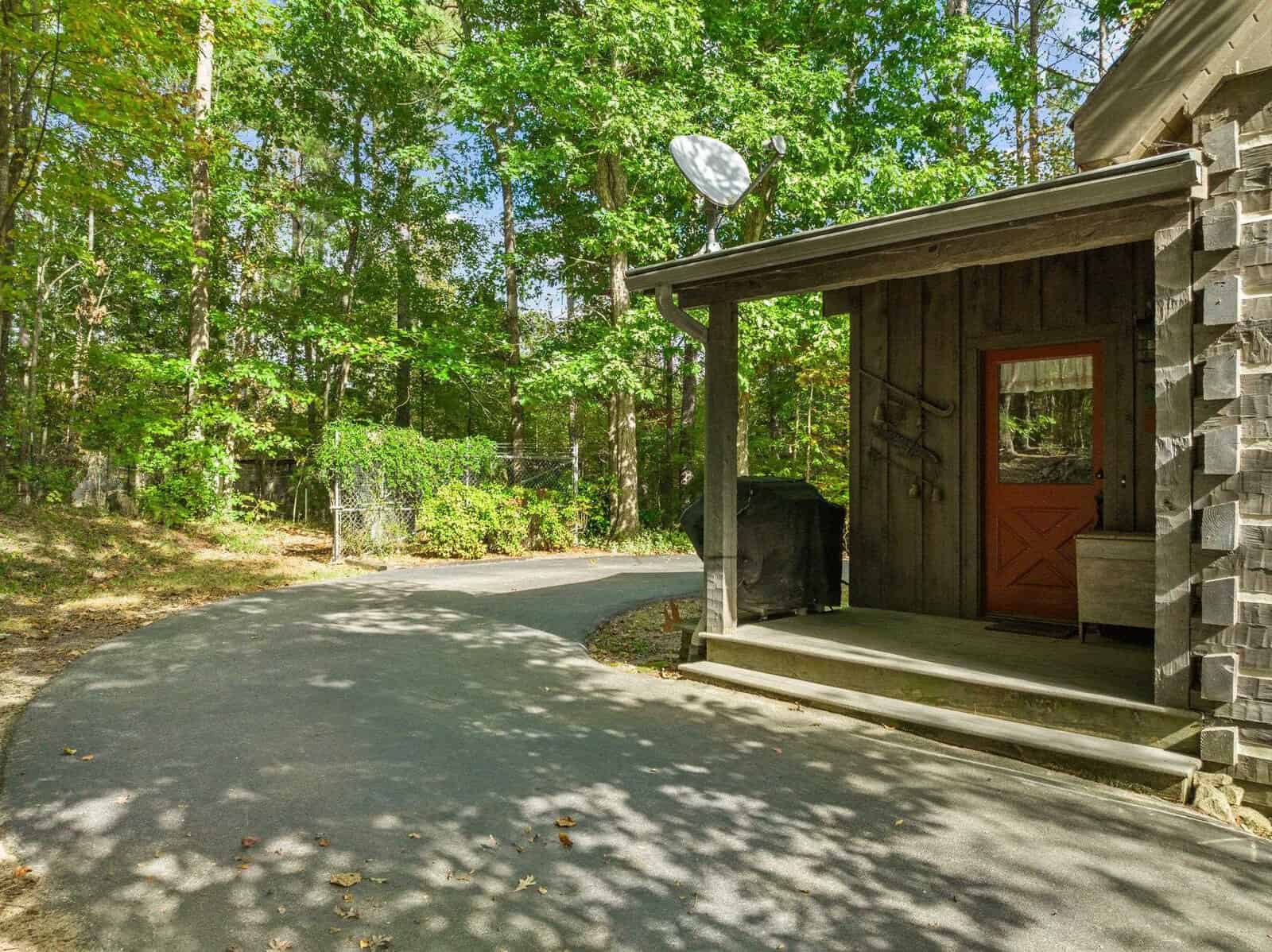 A paved driveway curves towards a rustic wooden building with a covered porch. A red door with glass panes is under the porch, which has a satellite dish on its roof. The area is surrounded by dense greenery, including trees and bushes.