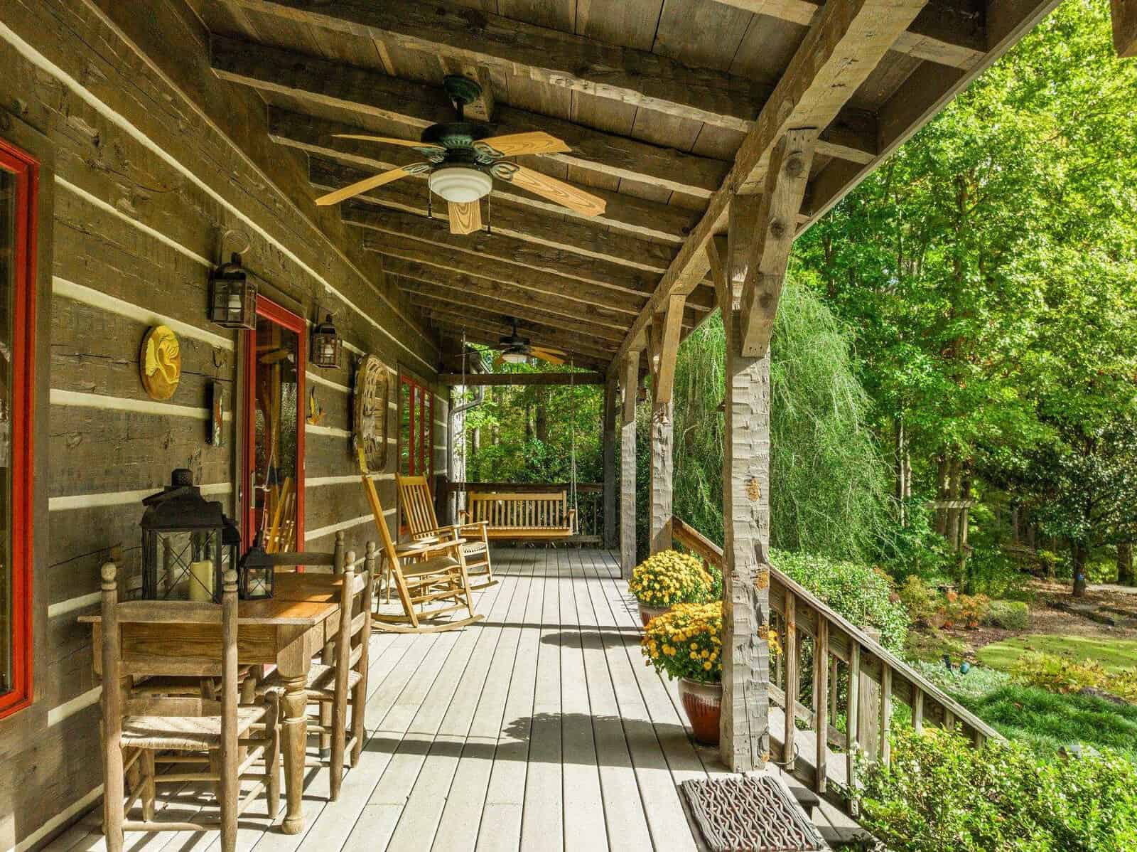 A rustic wooden porch with a ceiling fan, wooden chairs, a rocking chair, and a table with a lantern. Potted flowers are placed along the railing. Green trees and a grassy yard are visible in the background, bathed in sunlight.