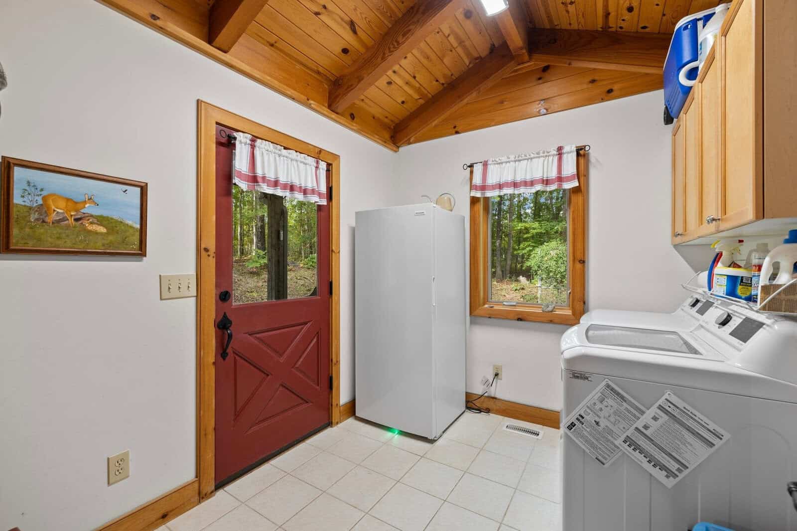 A laundry room with a wooden ceiling and a tiled floor. It features a red door with a window, a white refrigerator, a washing machine and dryer, cabinets, and shelves with laundry supplies. A picture of deer is hanging on the wall.