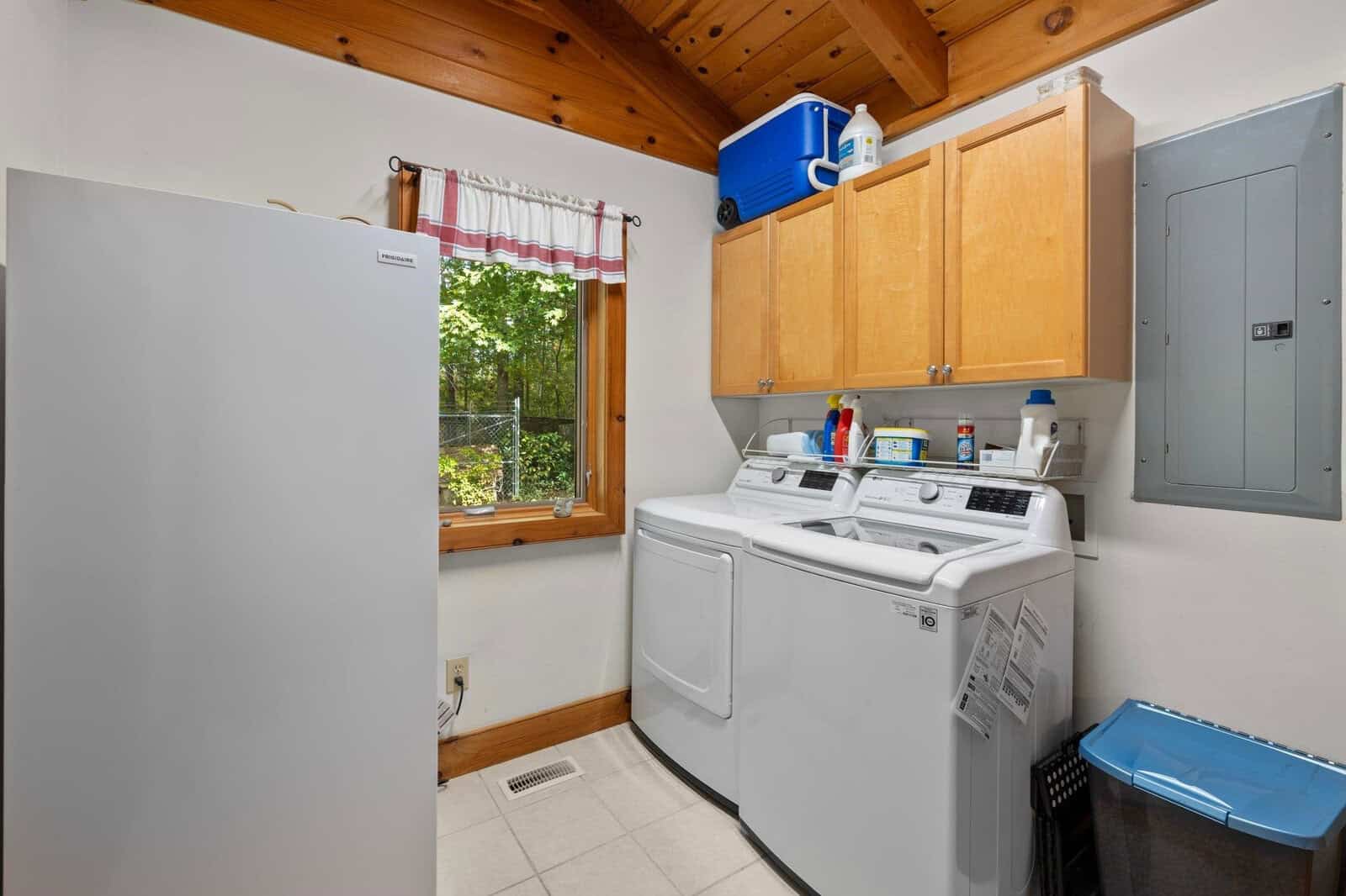 A laundry room with a washing machine and dryer side by side under a wooden cabinet. A fridge stands to the left, and various cleaning products are on the cabinet. A window with a curtain is on the back wall, revealing a green outdoor area.