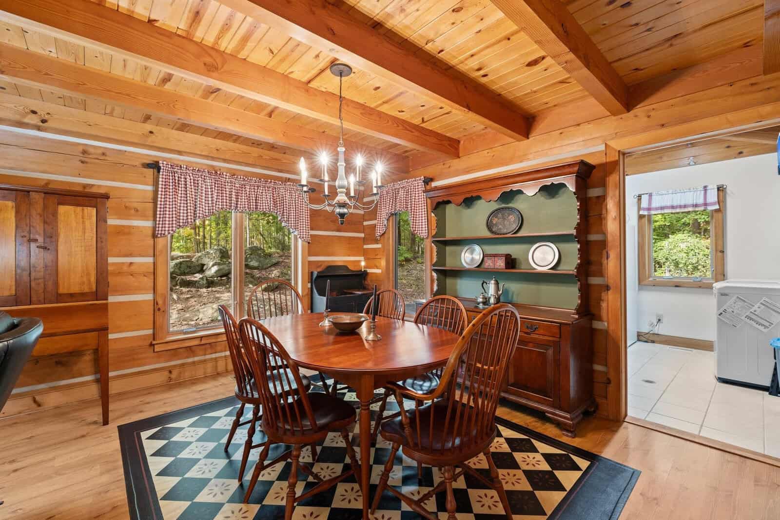 A cozy dining room features a wooden table with six matching chairs. The space is adorned with wood paneling and a beamed ceiling. A chandelier hangs above the table, and large windows with red checkered curtains allow natural light to fill the room.
