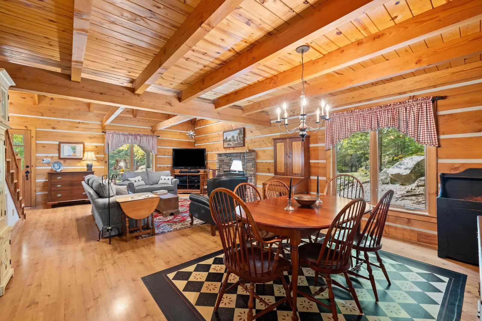A cozy log cabin interior featuring a dining area with a wooden table and six chairs, a living space with a couch and television, large windows with checkered curtains, hardwood floors, and exposed wooden beams. Natural light filters through the windows.