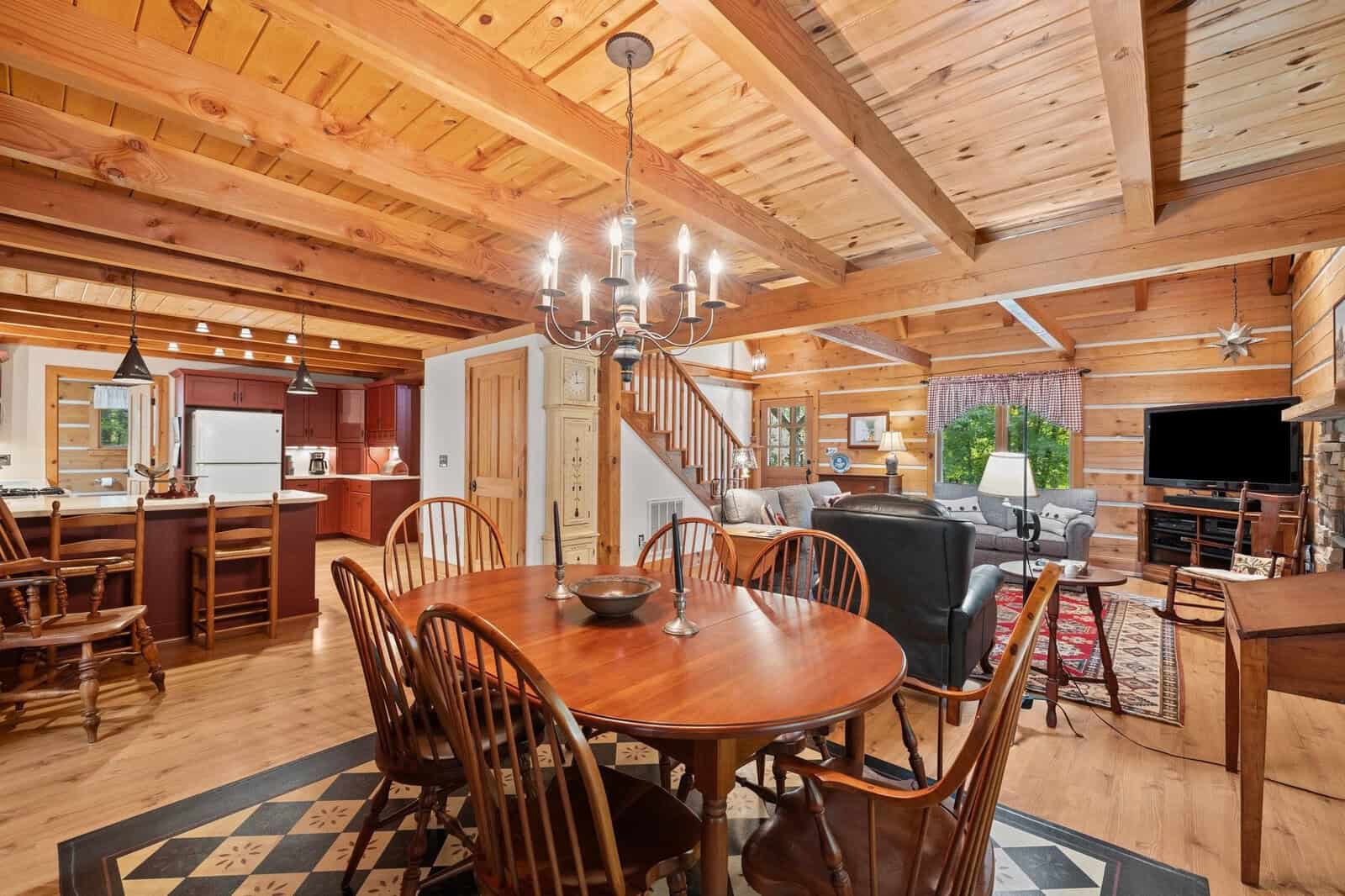 A cozy rustic dining room with a wooden table and six chairs is situated in an open-plan space with a visible kitchen and living area. Wooden beams adorn the ceiling, and a staircase leads to the upper floor. The space is warmly lit and features a mix of modern and vintage decor.