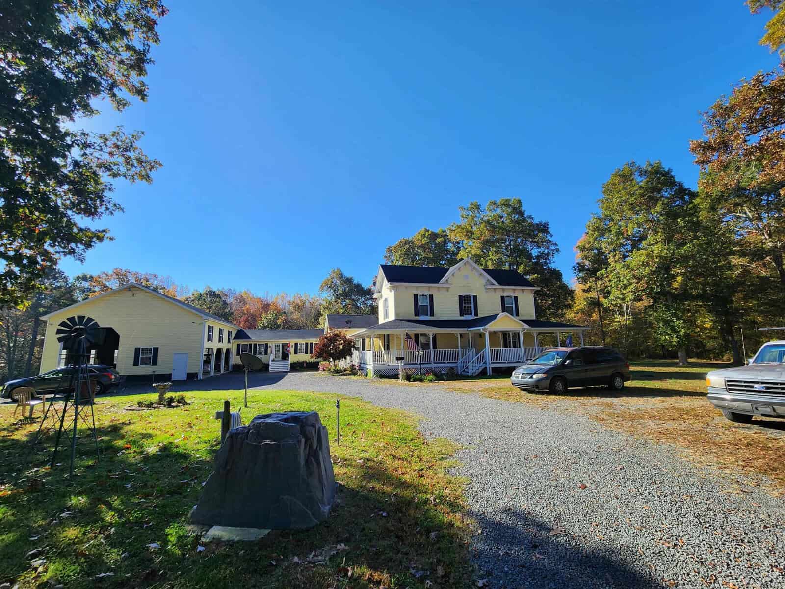 A sunny day reveals a large white house with a wraparound porch at 1170 Rocky Top Road. Cars are parked in the gravel driveway, while trees with autumn foliage surround the property. A large rock stands prominently in the foreground, adding to the rustic charm of this idyllic setting.