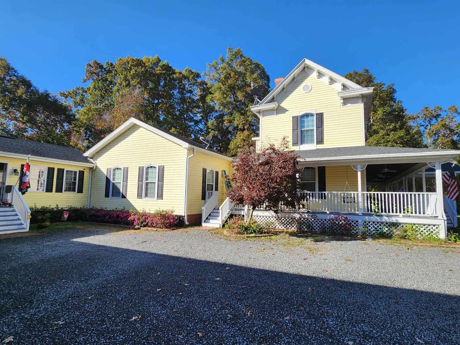 A two-story yellow house with a porch and two sections. The house has white trim and green shutters. A gravel driveway leads up to it, surrounded by colorful flowers and trees in the background under a clear blue sky.