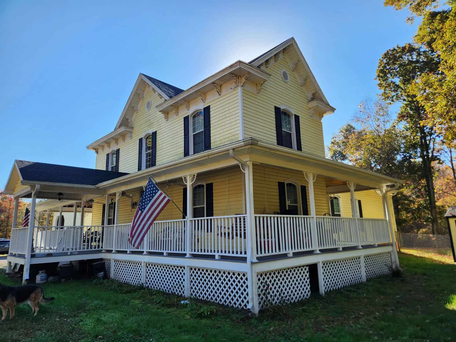 A yellow two-story house with a large wraparound porch and white trim. An American flag is displayed on the porch railing, and a dog is seen on the grass in the foreground. The sky is clear and sunny.
